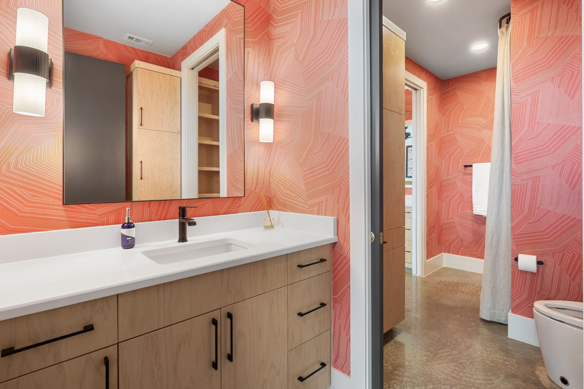 Modern bathroom with coral walls, wood vanity, white sink, and mirror, viewed toward toilet and shower area