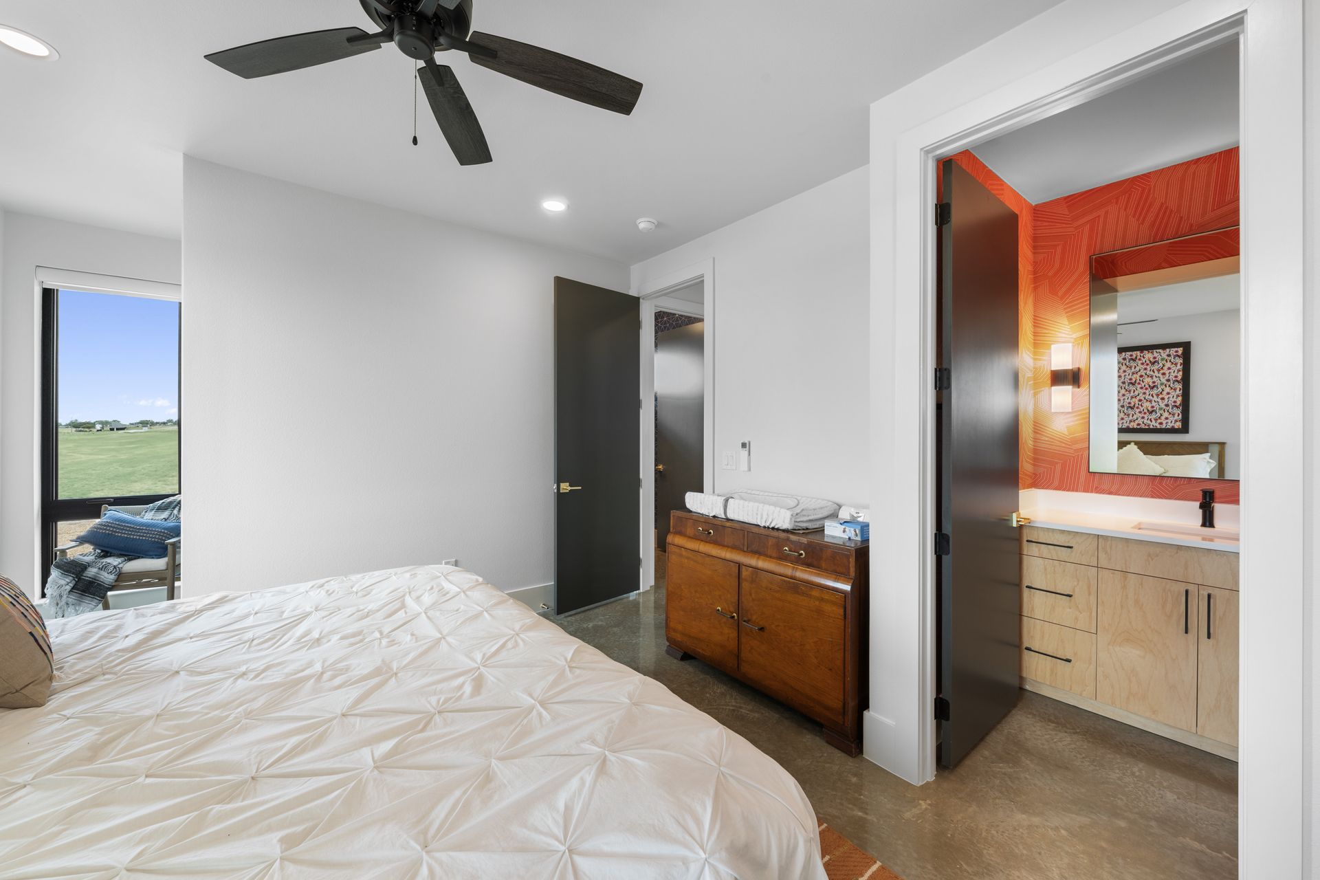 Bright bedroom with ceiling fan, open bathroom door, and window view of green fields.