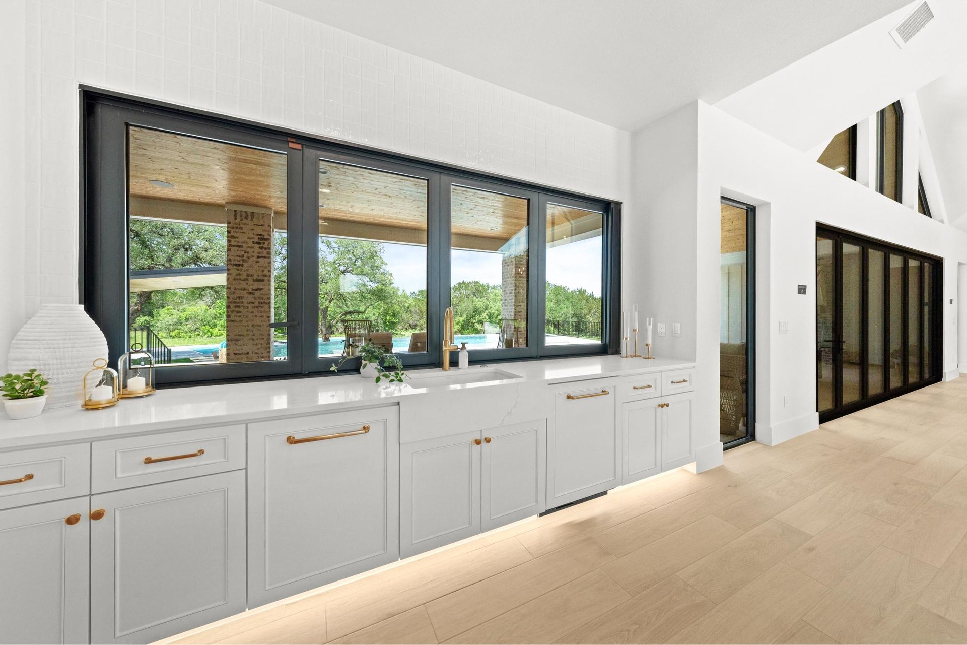 Bright white kitchen with long windows, gray cabinets, and light wood flooring overlooking greenery