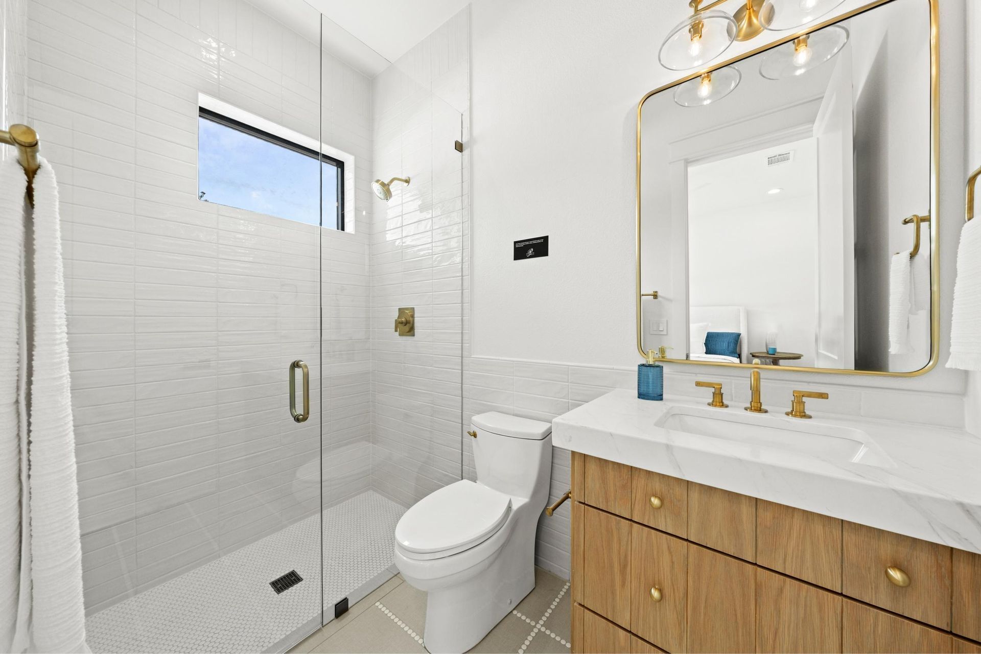 Bright white bathroom with glass shower, toilet, and wood vanity with gold fixtures