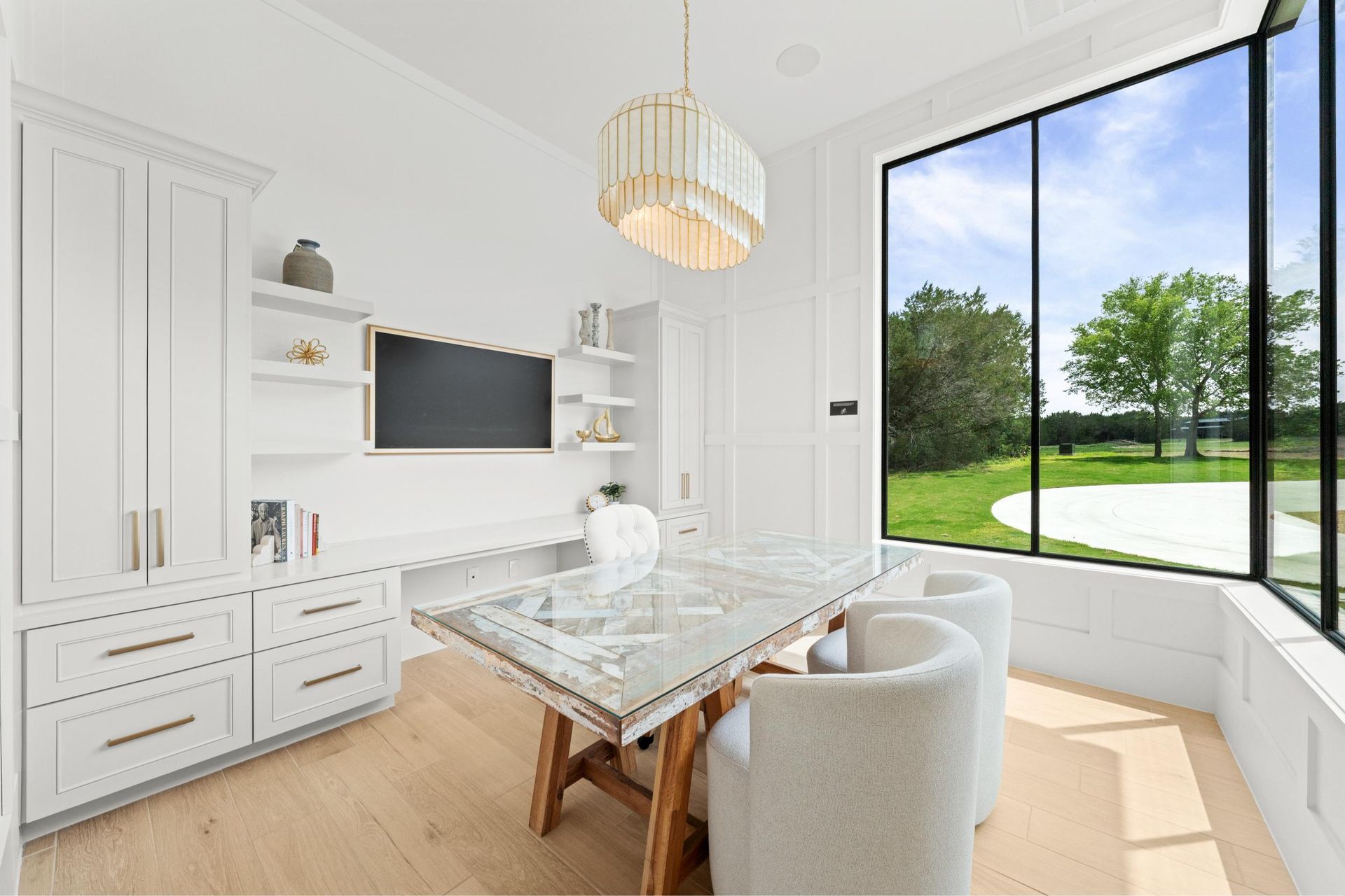 Bright white home office with built-in desks, a glass table, and large windows overlooking a green lawn