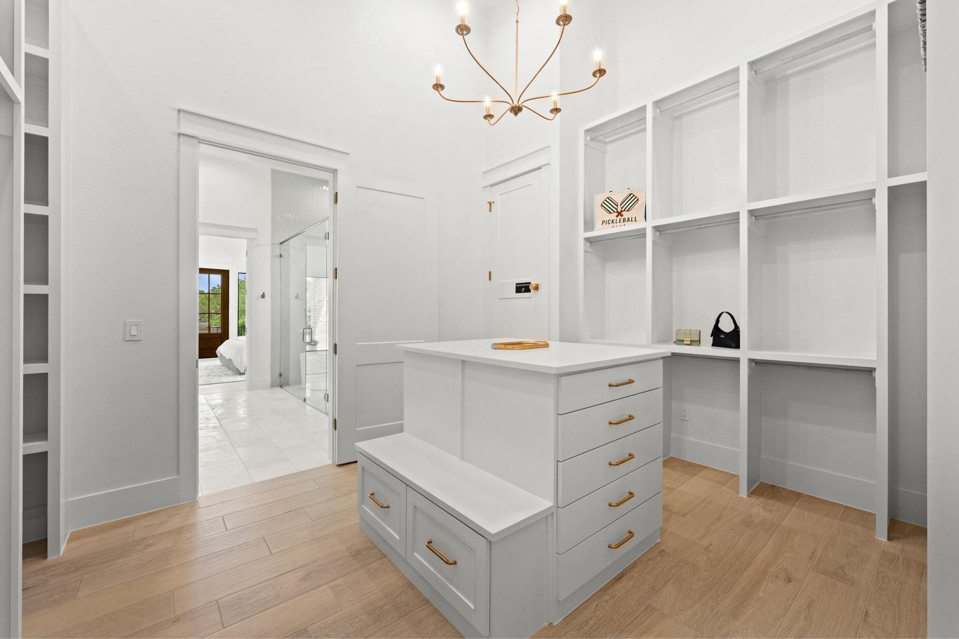 Bright white walk-in closet with built-in shelves, drawers, and a chandelier over a wood floor.