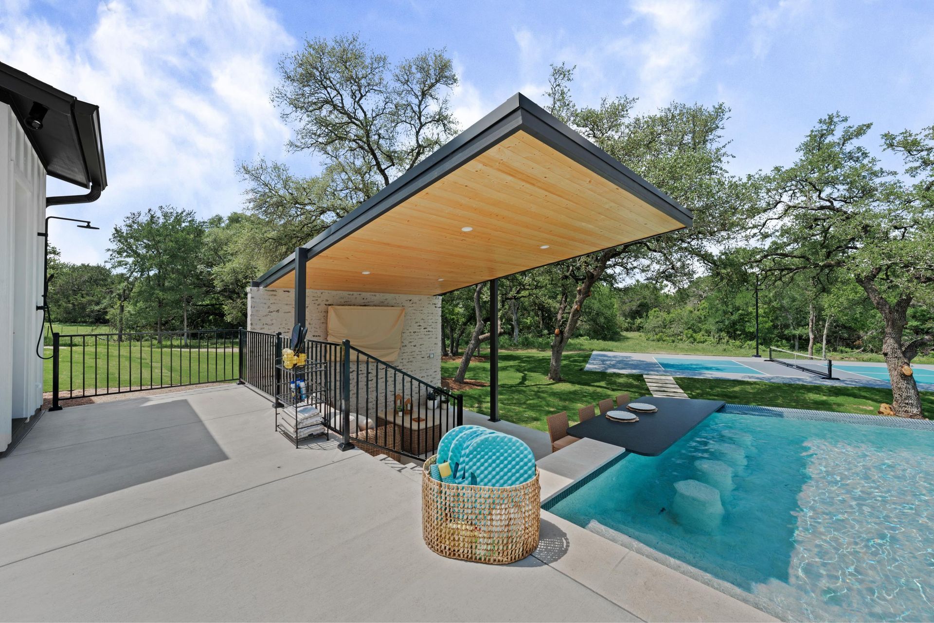 Covered patio beside a turquoise pool, with outdoor seating and trees overlooking the water