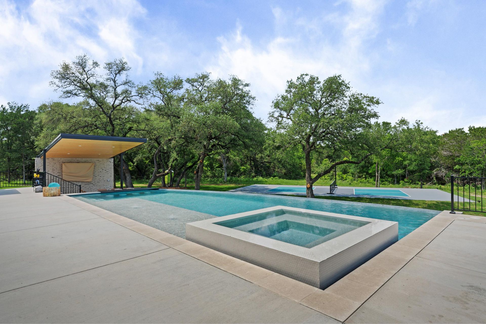 Modern outdoor pool with a square hot tub, patio, and trees under a blue sky