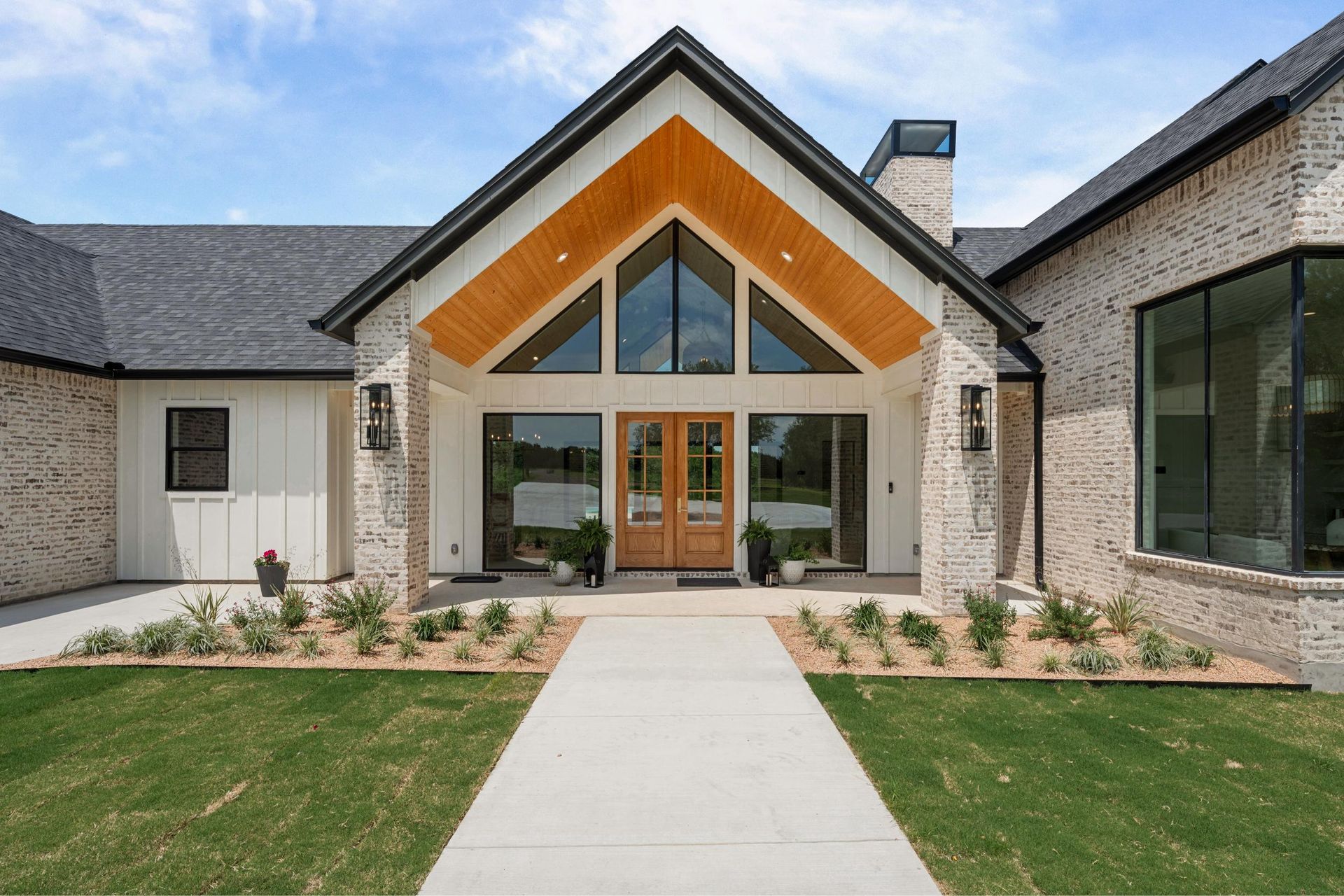 Modern stone house entrance with tall wooden doors, large windows, and a central walkway across a lawn