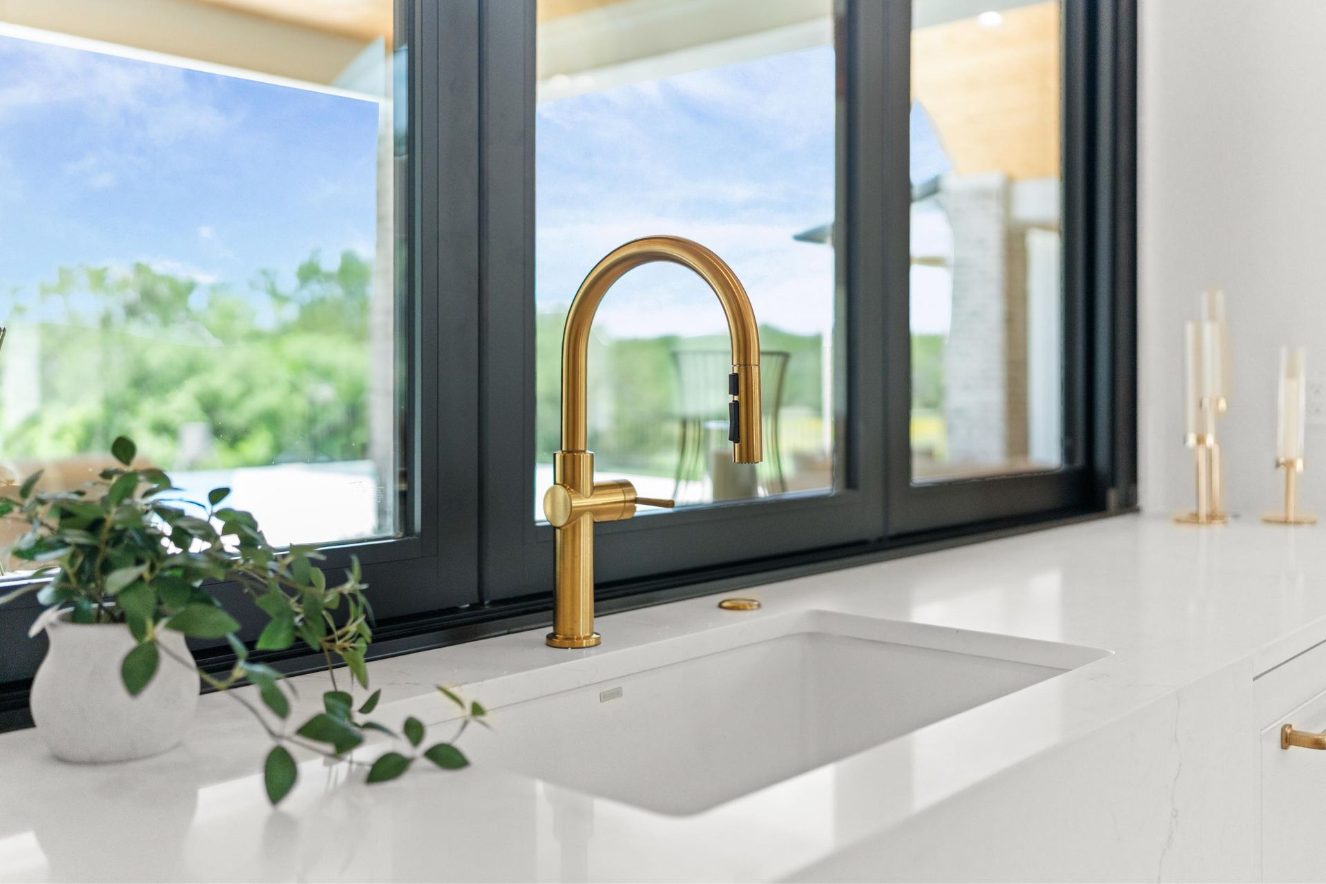 Modern kitchen sink with gold faucet beside a large window and a small potted plant