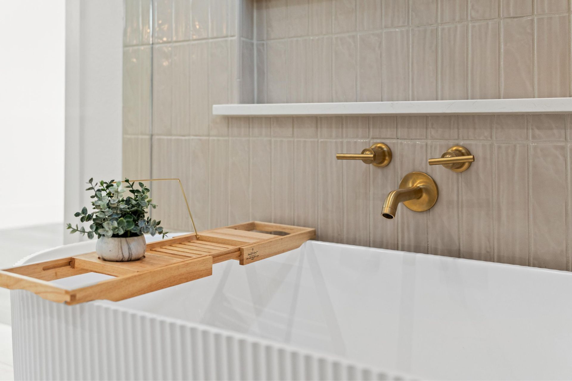 Wooden bath tray over a white tub beside gold faucet and beige tiled wall