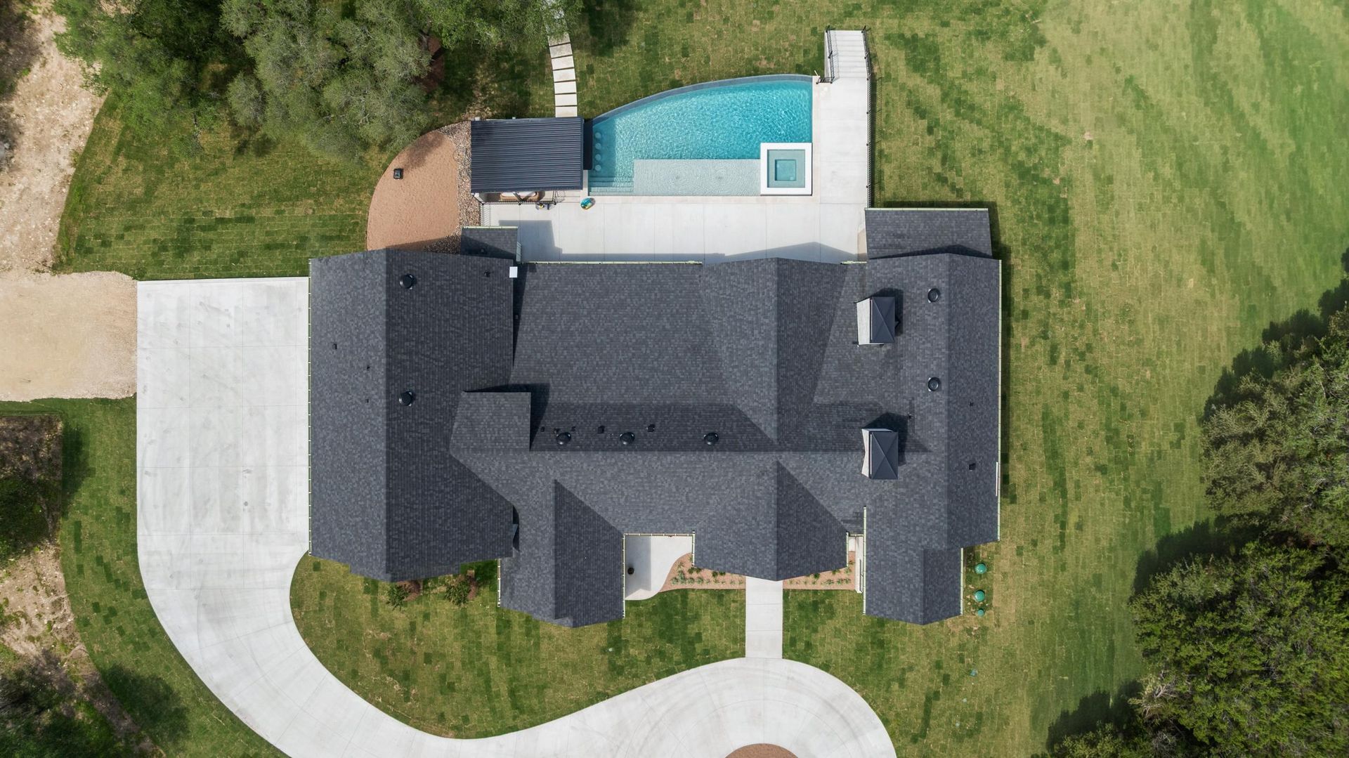 Aerial view of a house with a dark roof, curved driveway, and backyard swimming pool
