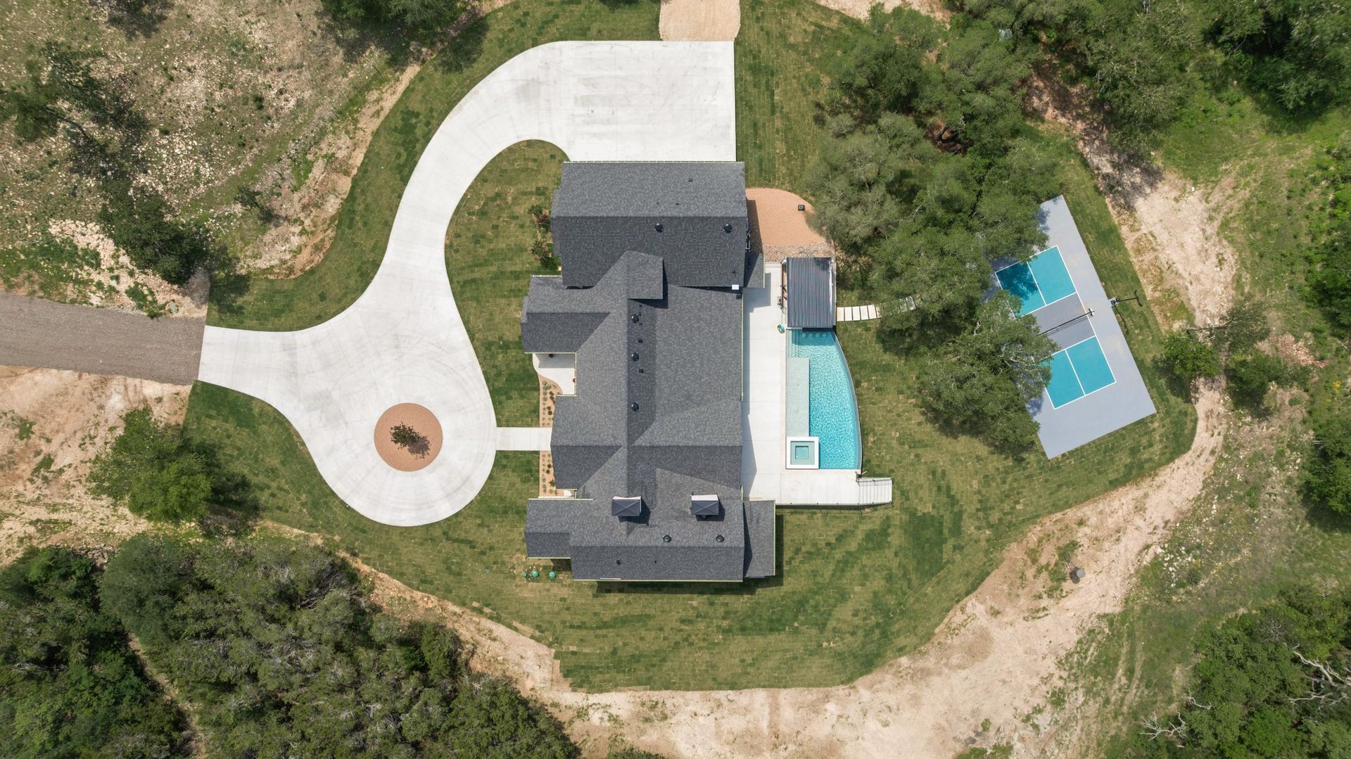 Aerial view of a large gray house with a curved driveway, patio, and blue swimming pool surrounded by trees.