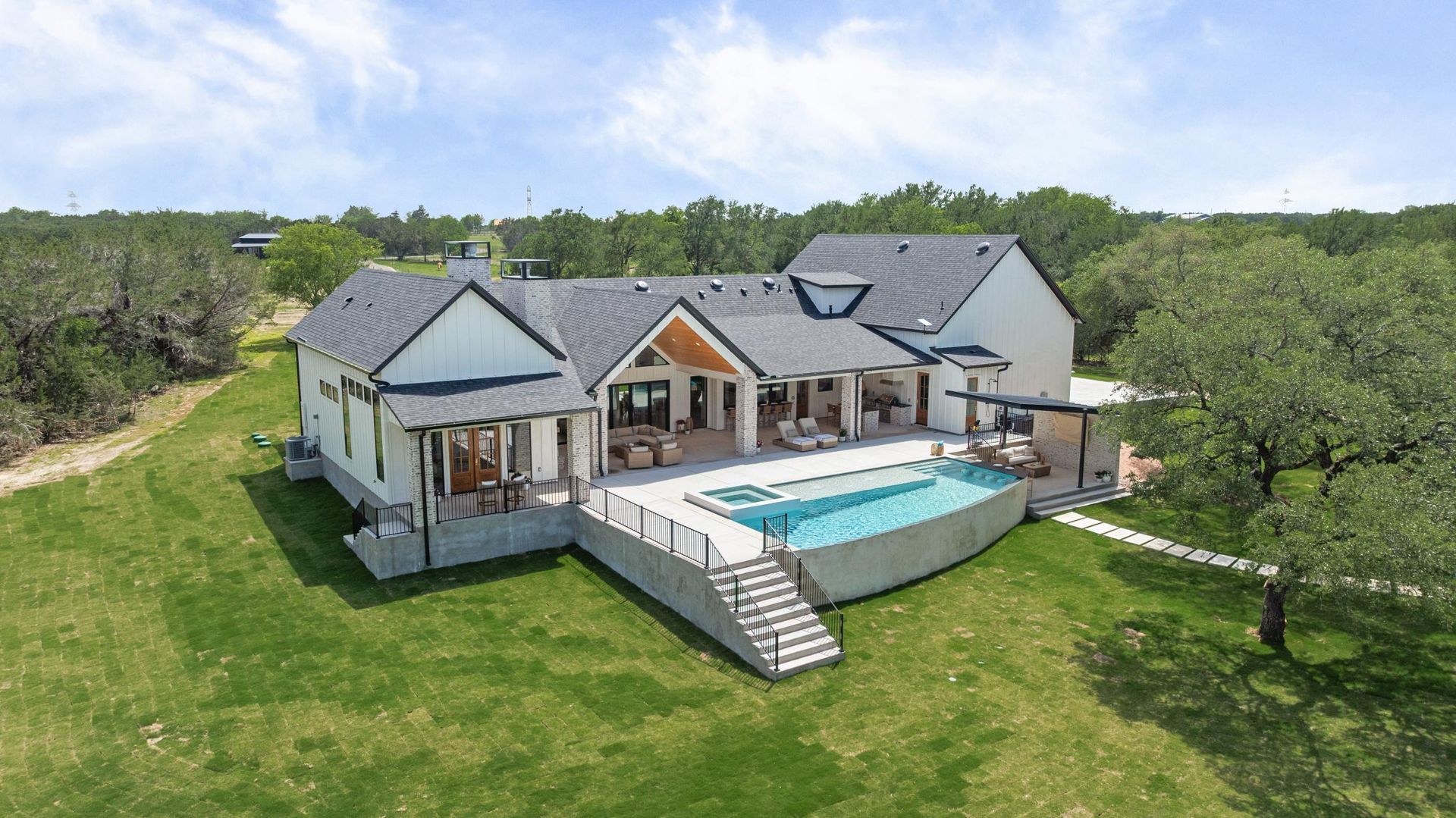 Modern house with a wraparound deck and pool on a grassy hillside, surrounded by trees