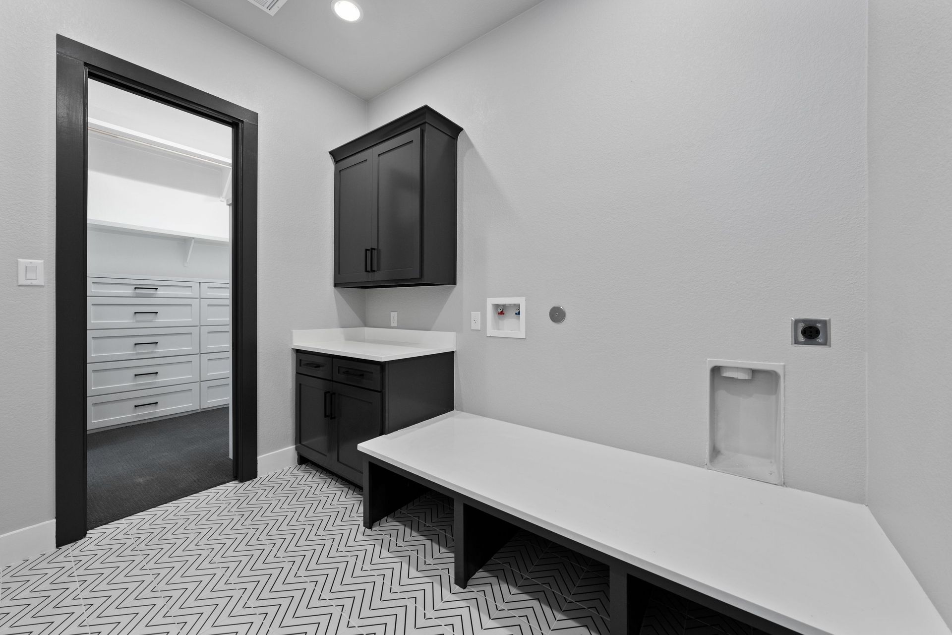 Modern laundry room with black cabinets, white counters, patterned floor, and a long worktable
