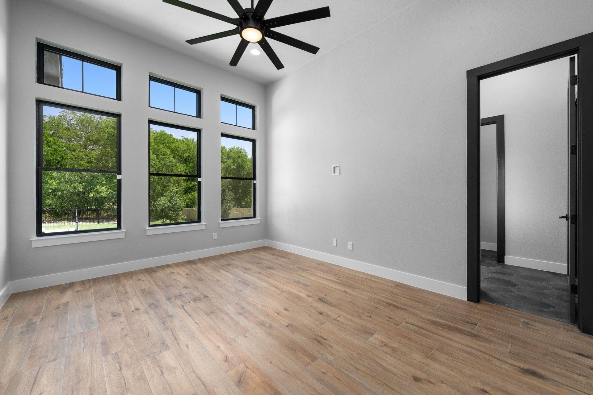 Bright empty room with large windows, light wood floor, and a black ceiling fan; open doorway on the right