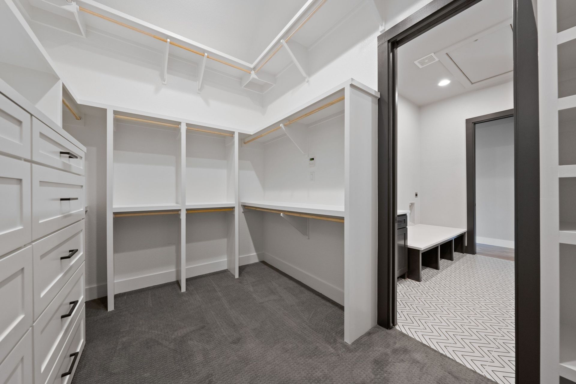 White walk-in closet with built-in shelves, hanging rods, drawers, and gray carpet, next to a tiled room