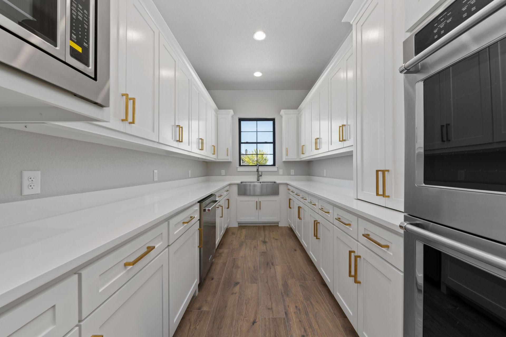 Bright modern galley kitchen with white cabinets, gold handles, wood floor, and a window at the end.