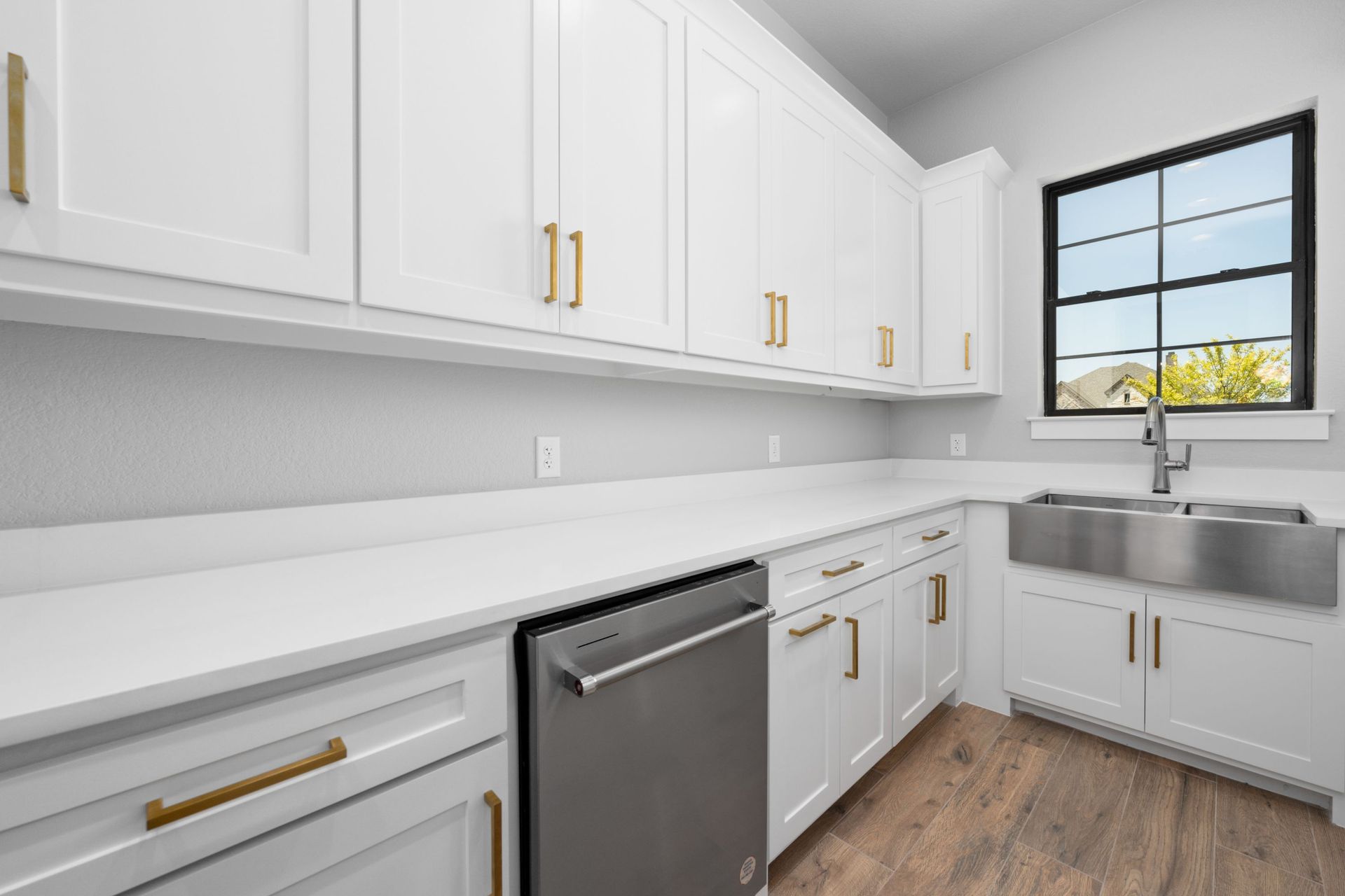 Bright white kitchen with gold handles, stainless dishwasher, and large window over the sink.