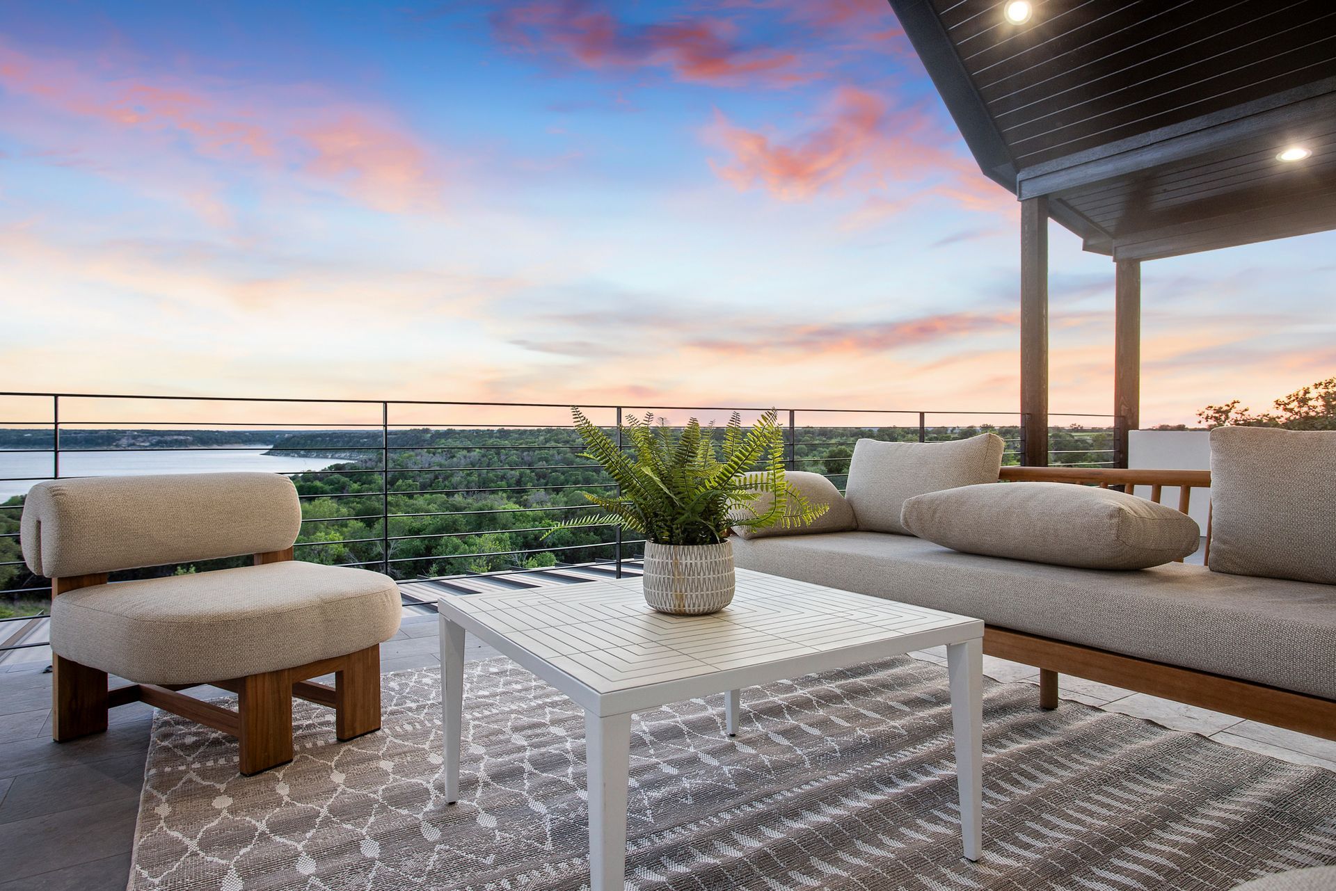 Modern patio with beige lounge chairs, white table, potted plant, and sunset ocean view.