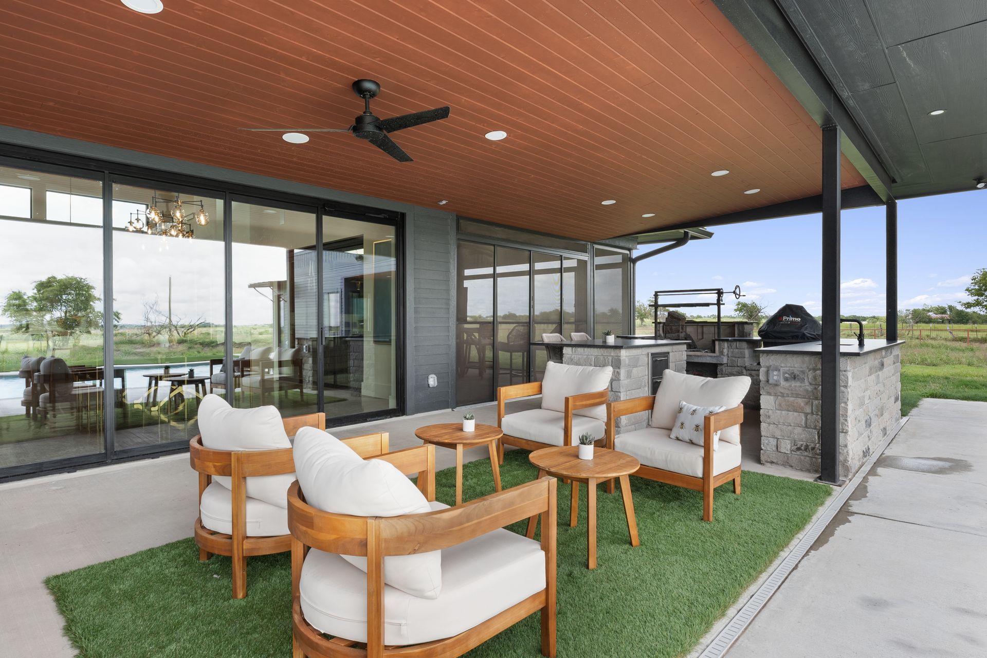Covered patio seating area with white-cushioned chairs, wooden tables, and glass walls by a building