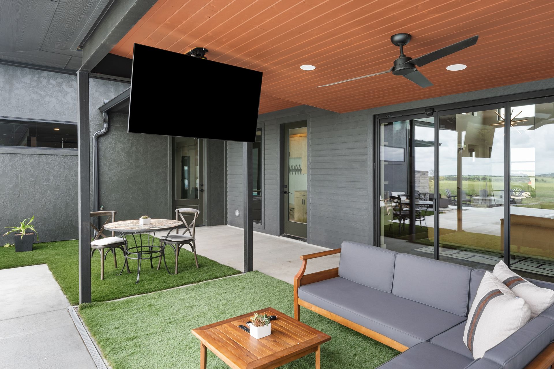 Covered patio with gray sofa, wooden table, dining set, and hanging TV beside sliding glass doors