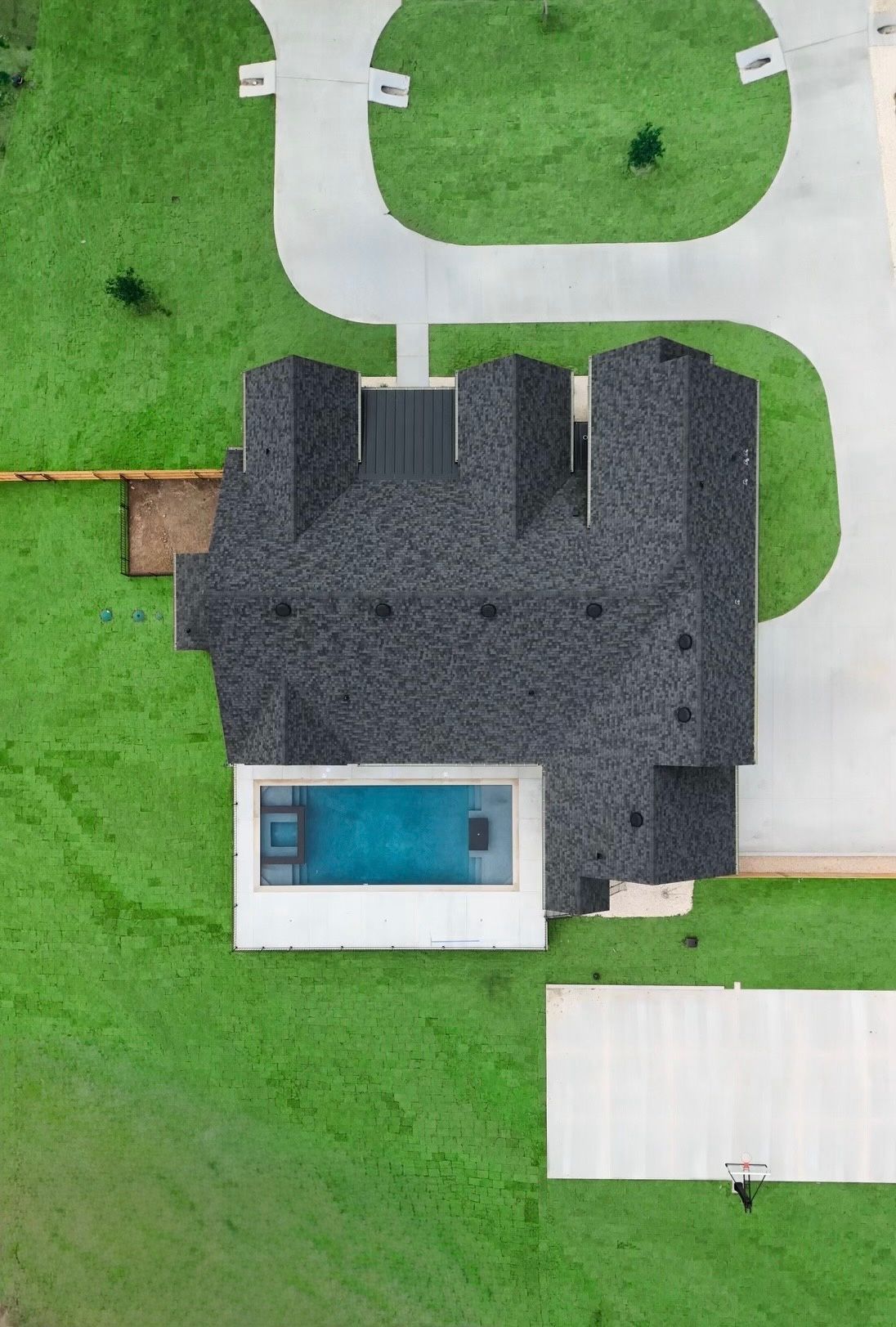Aerial view of a house with a dark roof, backyard pool, and surrounding green lawn.