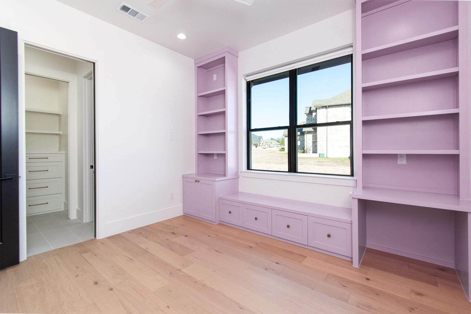 Empty lavender bedroom with built-in shelves, window seat, and light wood floor