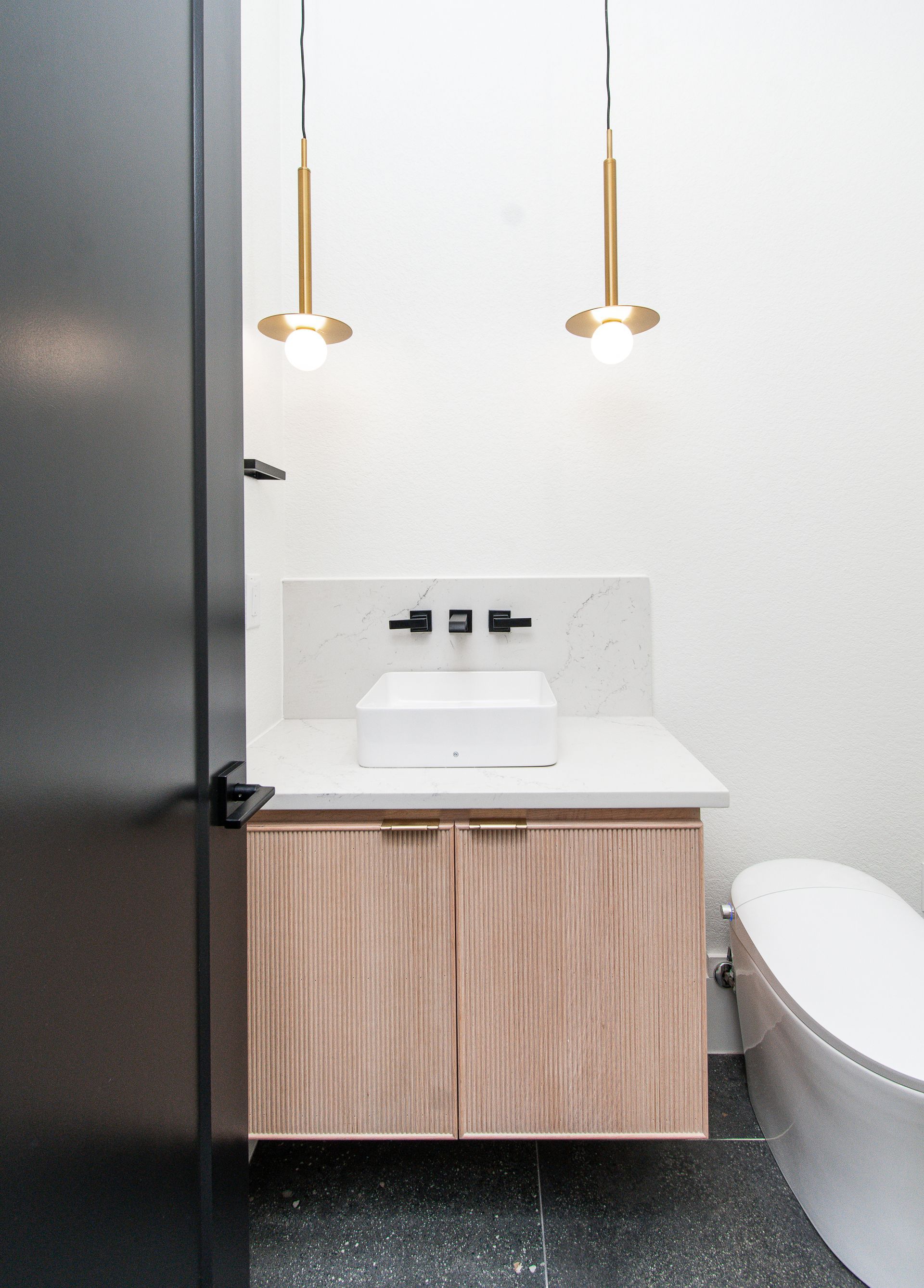 Small modern bathroom with a floating wood vanity, round sink, pendant lights, and a toilet seat visible on the right.