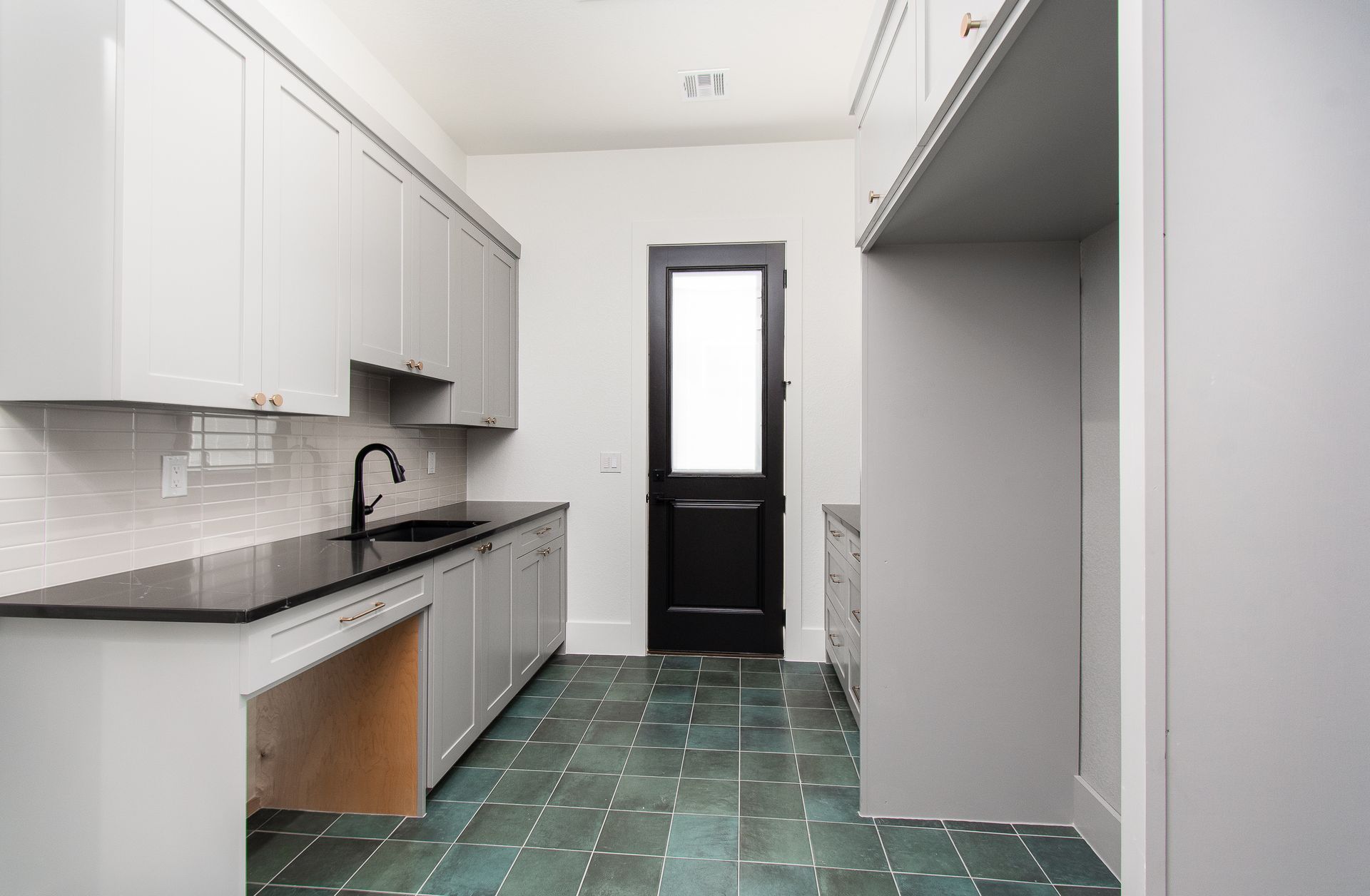 Narrow kitchen with white cabinets, black countertops, green tile floor, and a door at the far end.