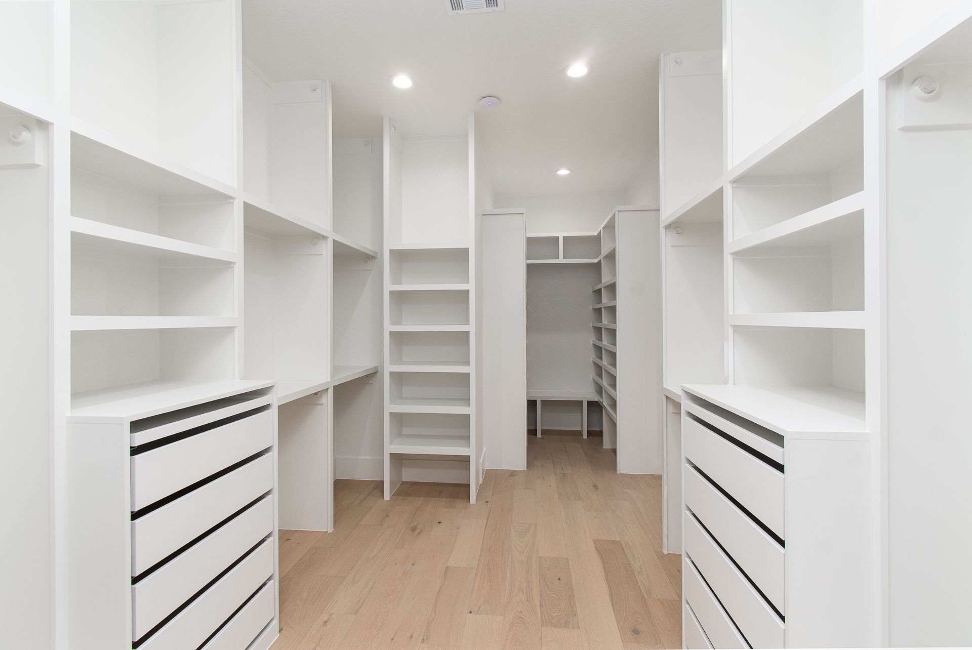 Bright white walk-in closet with built-in shelves and drawers on both sides, light wood floor, and recessed lighting