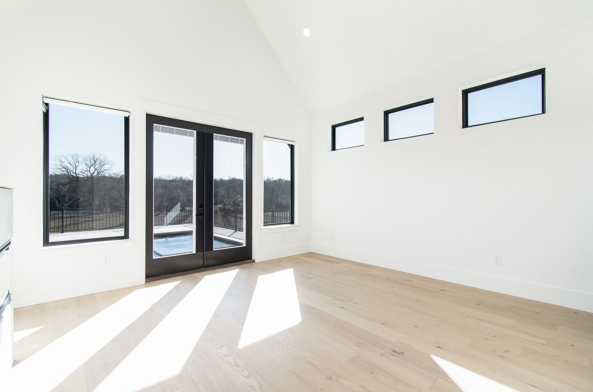 Bright empty room with white walls, light wood floor, black-framed windows, and sunlight streaming in