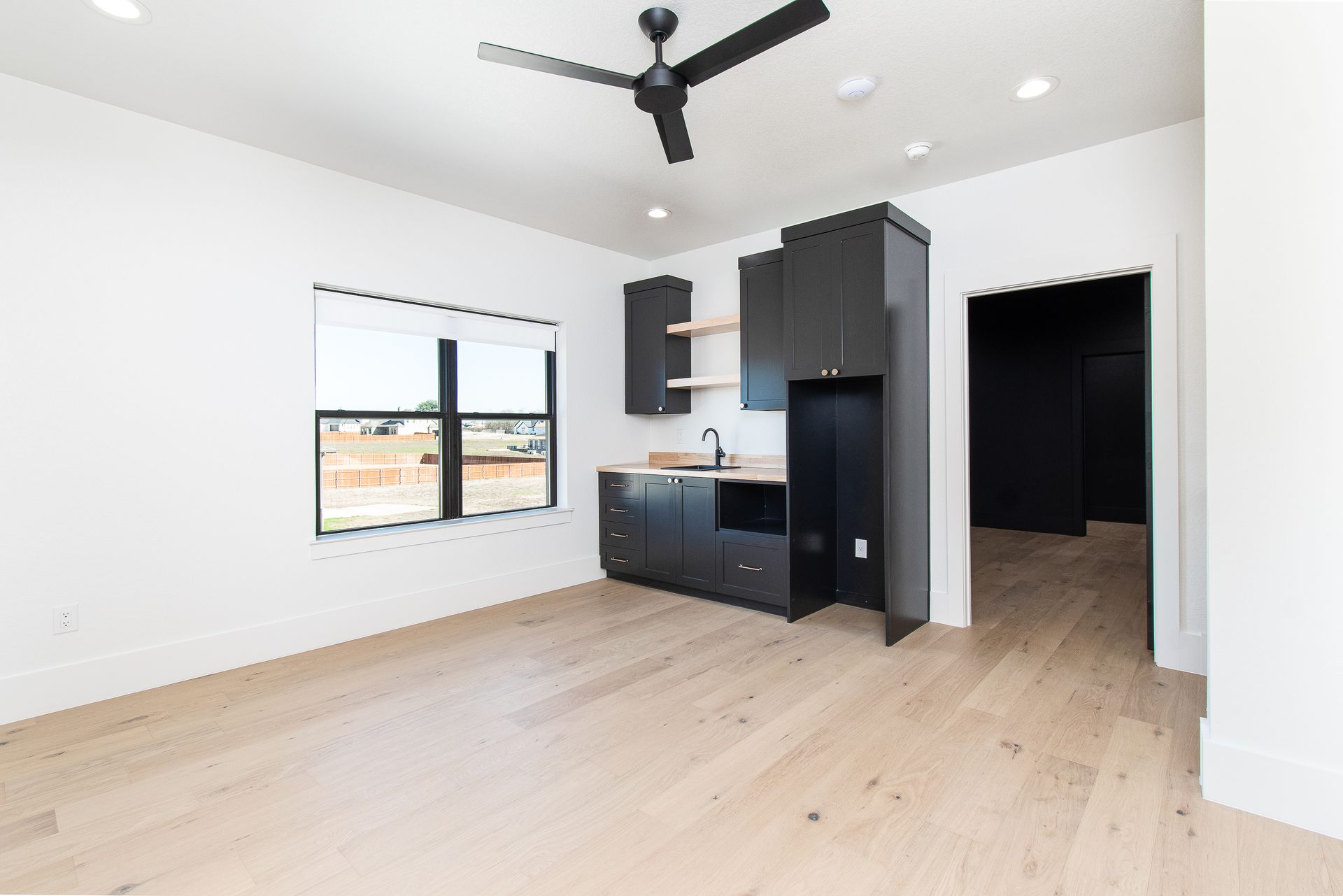 Empty modern apartment room with light wood floors, black kitchen cabinets, and a ceiling fan