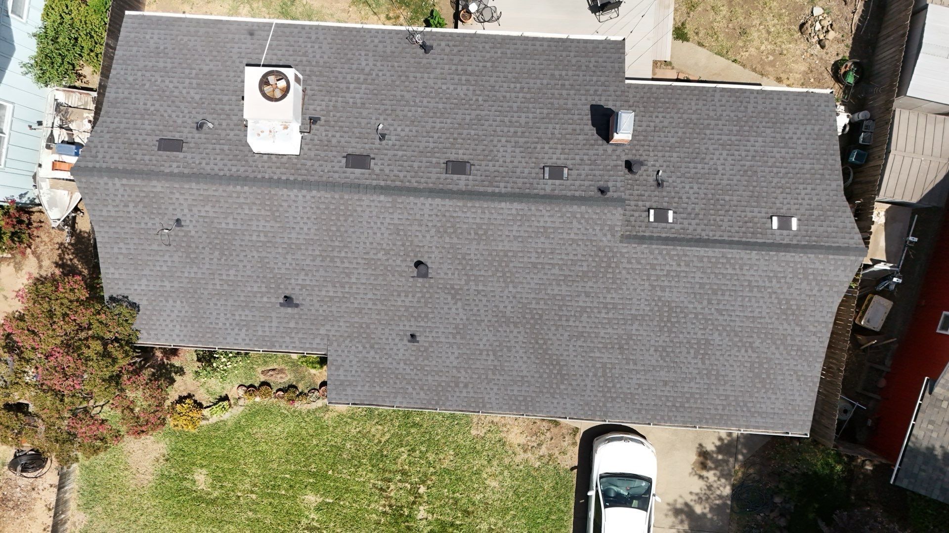 Aerial view of a gray-shingled house roof with a chimney, surrounding yard, and a parked white car nearby.