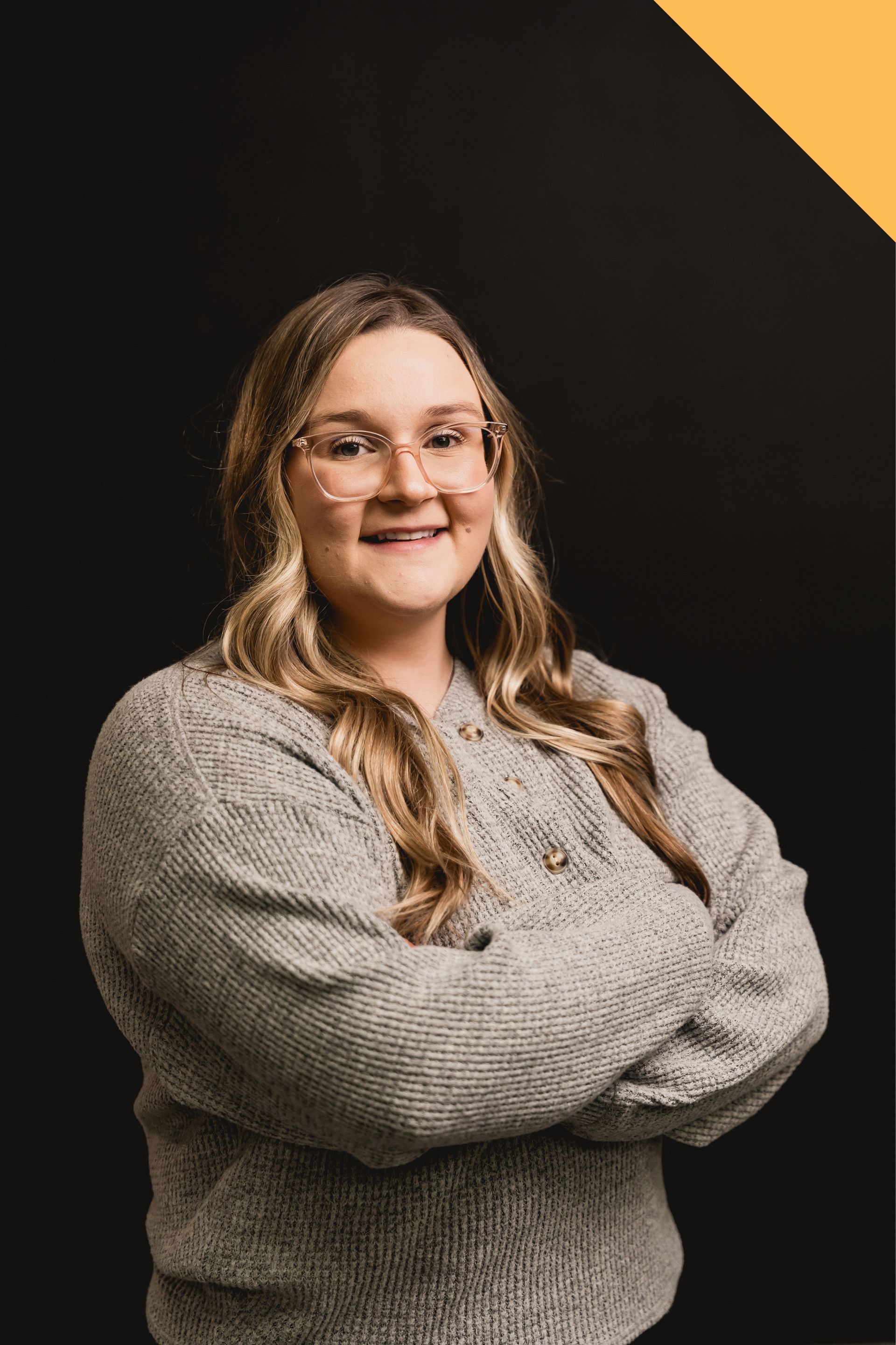 Person with glasses and long hair, arms crossed, wearing a gray sweater against a dark background with a yellow corner accent
