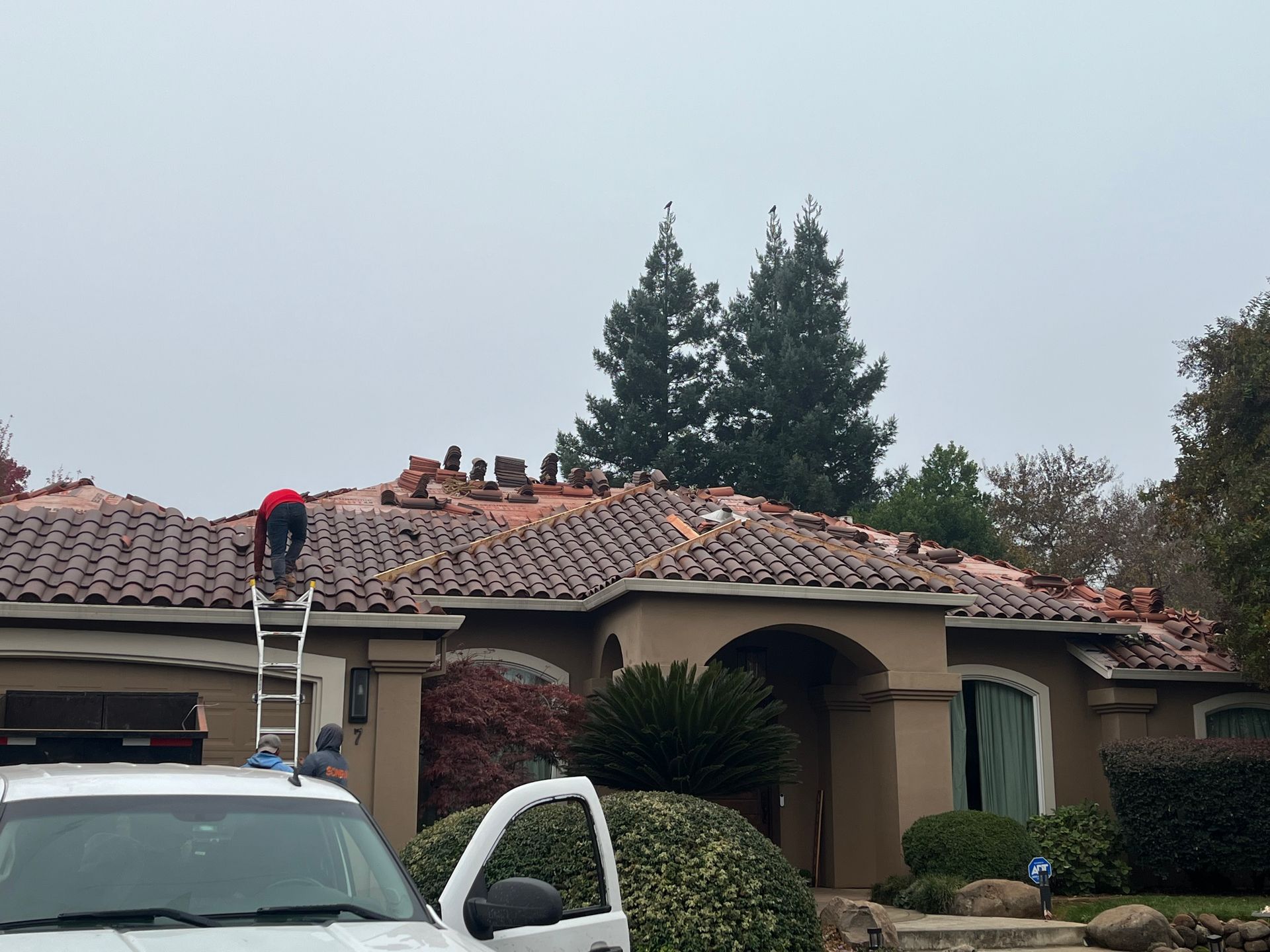 Worker on a ladder repairing a red-tiled roof on a suburban house, with a car parked in front.