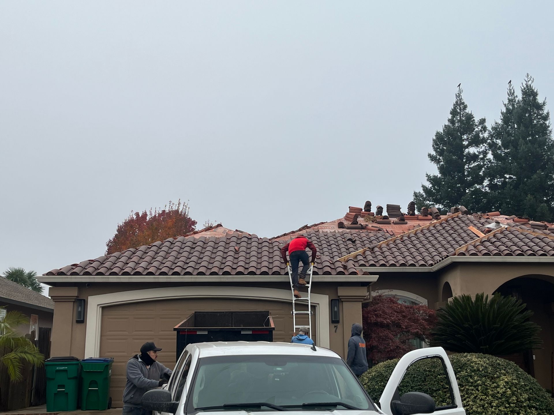 Worker on ladder on a house roof while another person stands by a white truck in a driveway.
