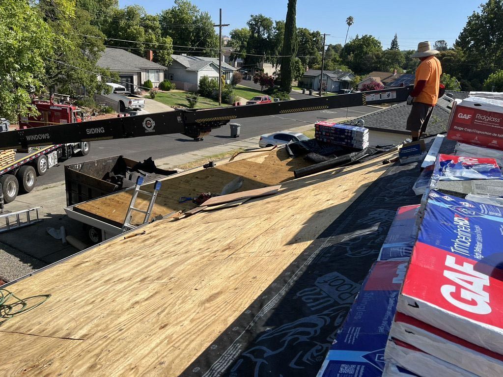 Roofer on a sloped roof installing plywood sheathing beside stacks of roofing materials on a sunny street.