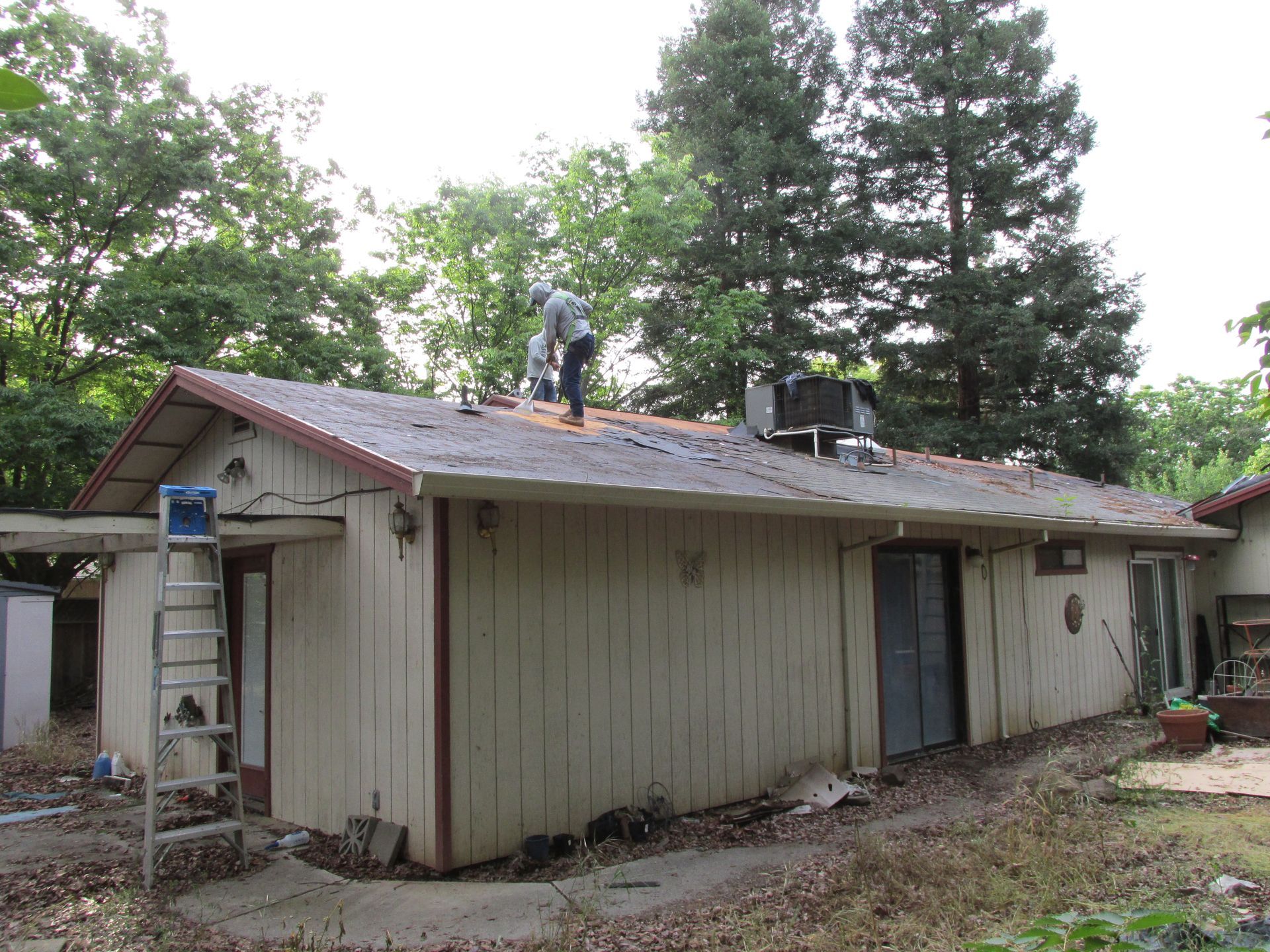 Workers repairing a roof on a beige house with a ladder and trees in the yard