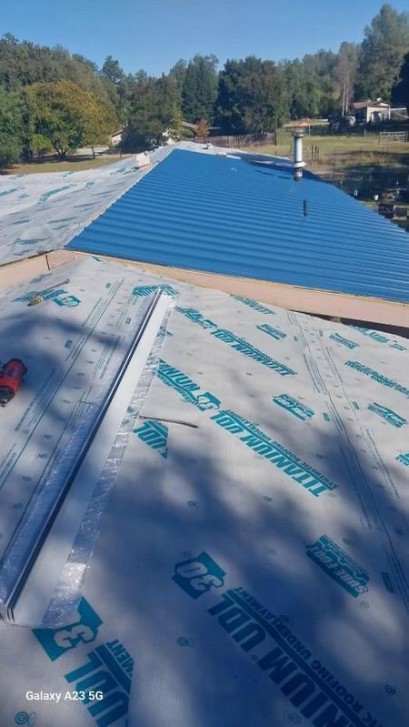 Blue metal roof under construction with protective underlayment, surrounded by trees and a clear sky