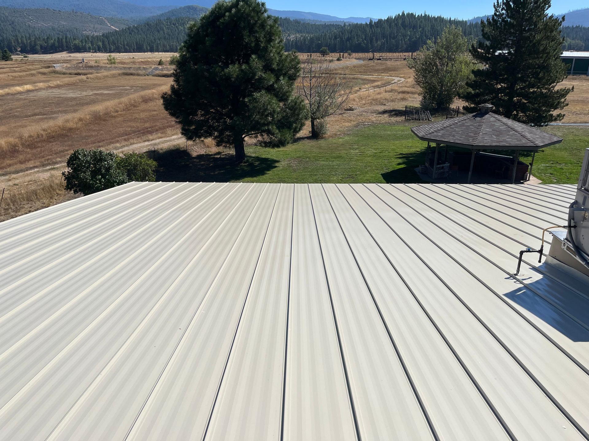 White metal roof overlooking a dry field, trees, and distant mountains under a clear sky