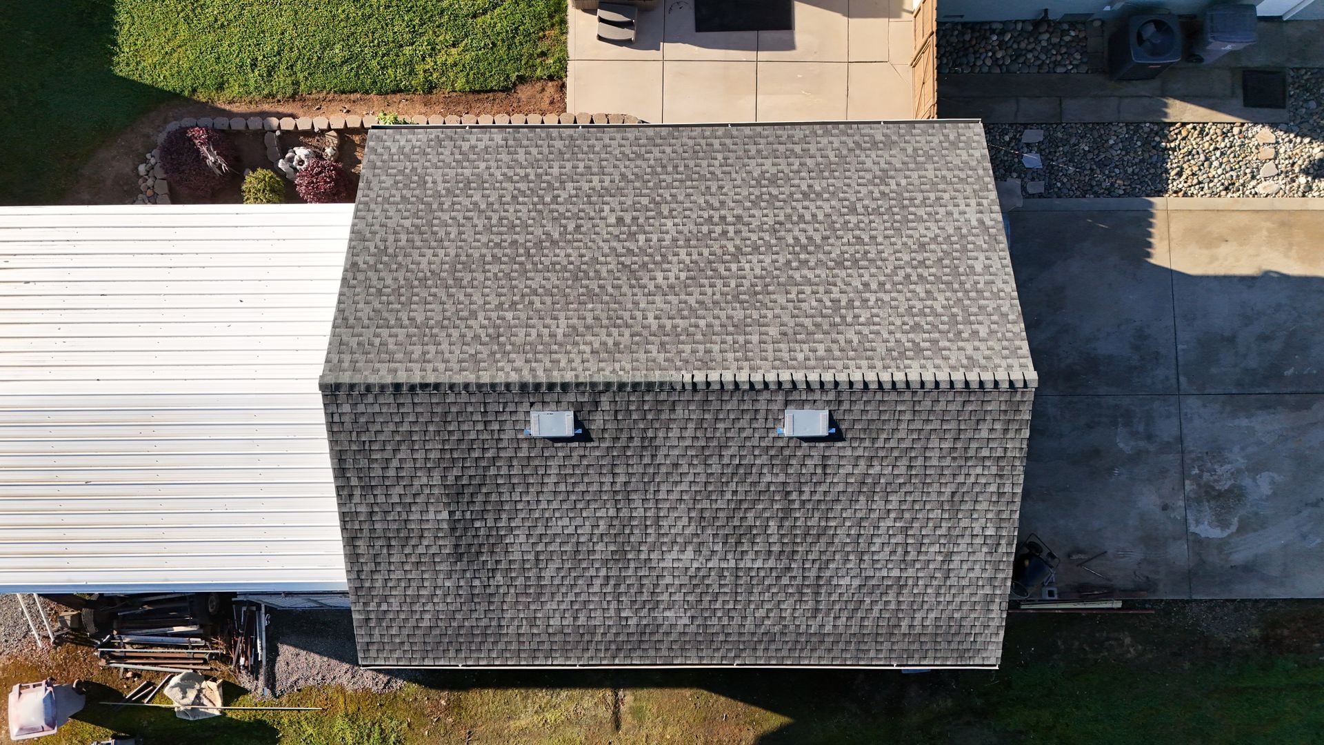 Aerial view of a gray-shingled roof beside a white-roofed structure and concrete driveway.