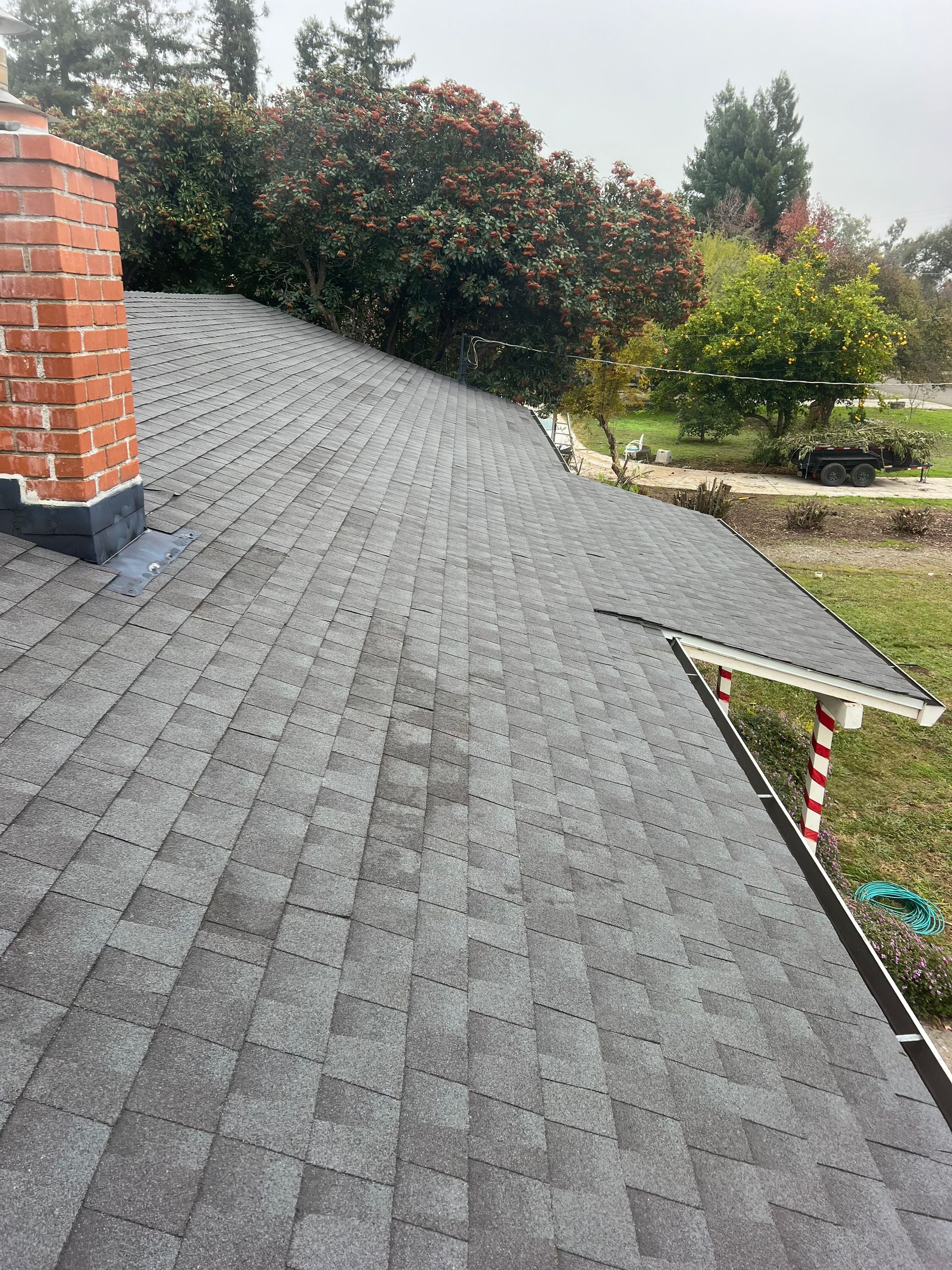 Gray shingled roof with brick chimney and a small extension, viewed from above amid trees and a yard