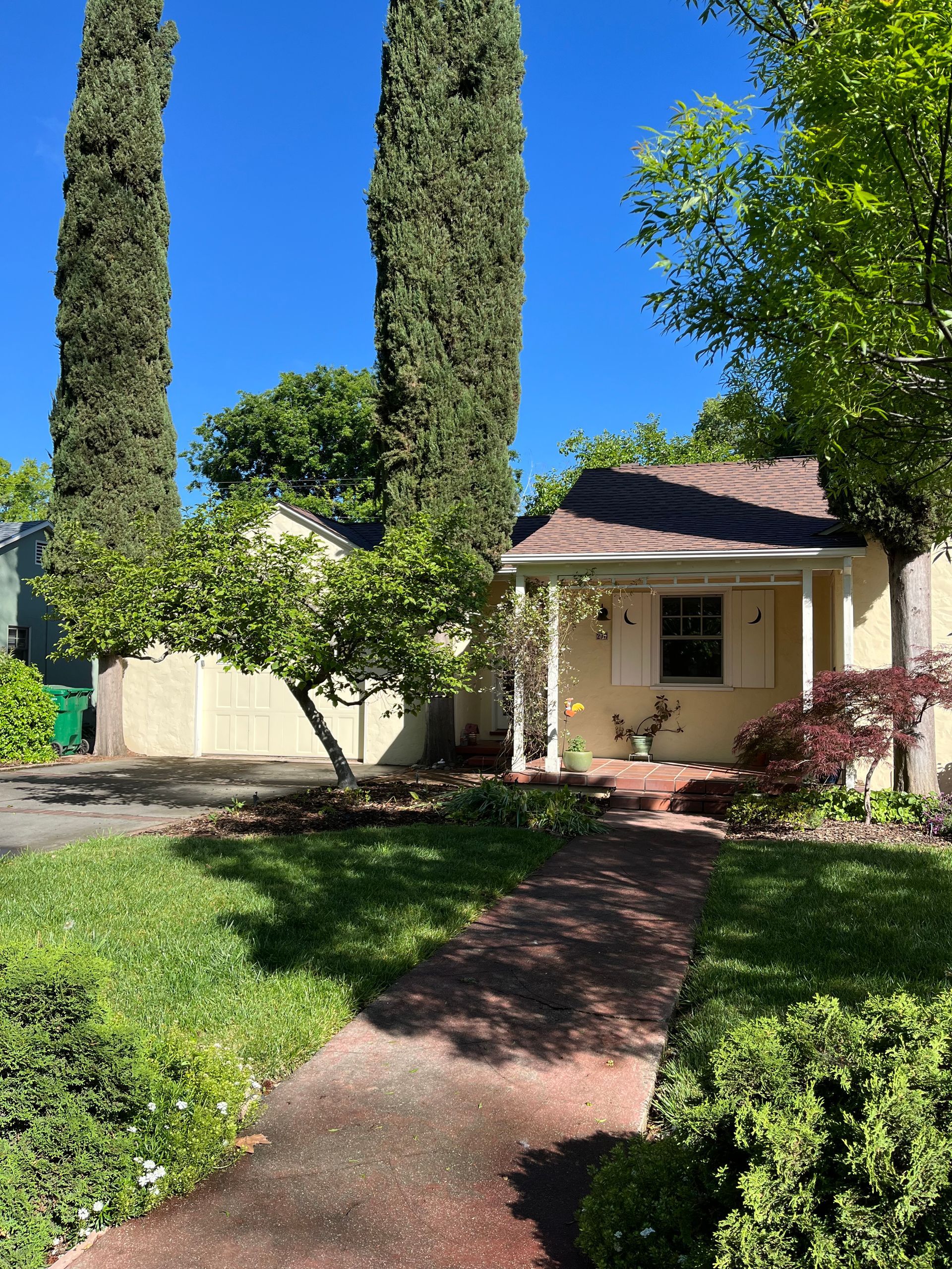 Small cream house with red roof, front path, green lawn, and tall cypress trees under a bright blue sky.