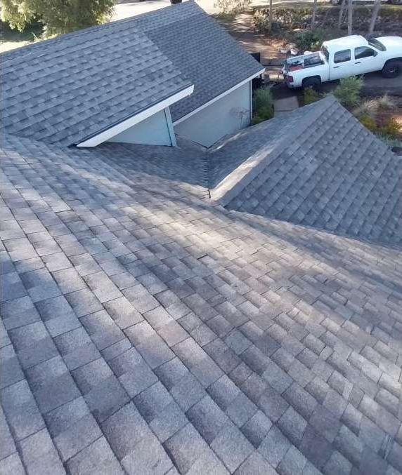 Gray shingled residential roof with a dormer and two white pickup trucks in a driveway below
