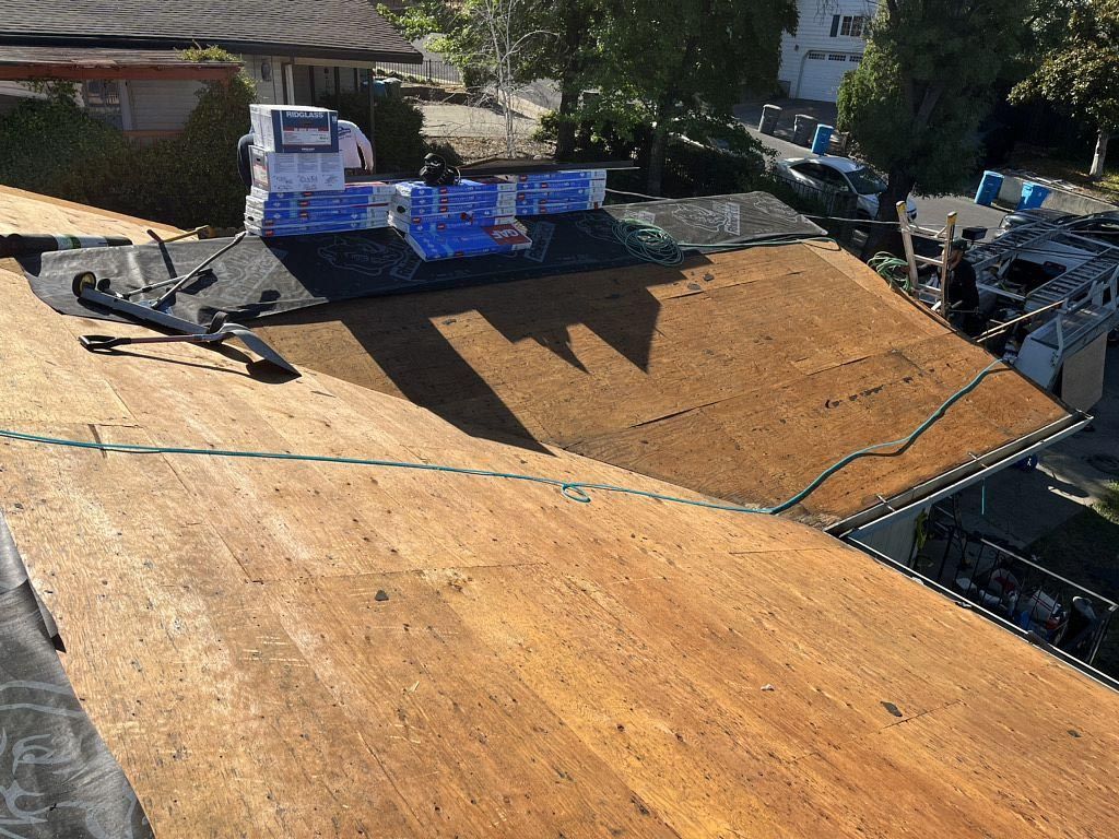 Roof under construction with plywood sheathing, workers, and stacked blue materials nearby