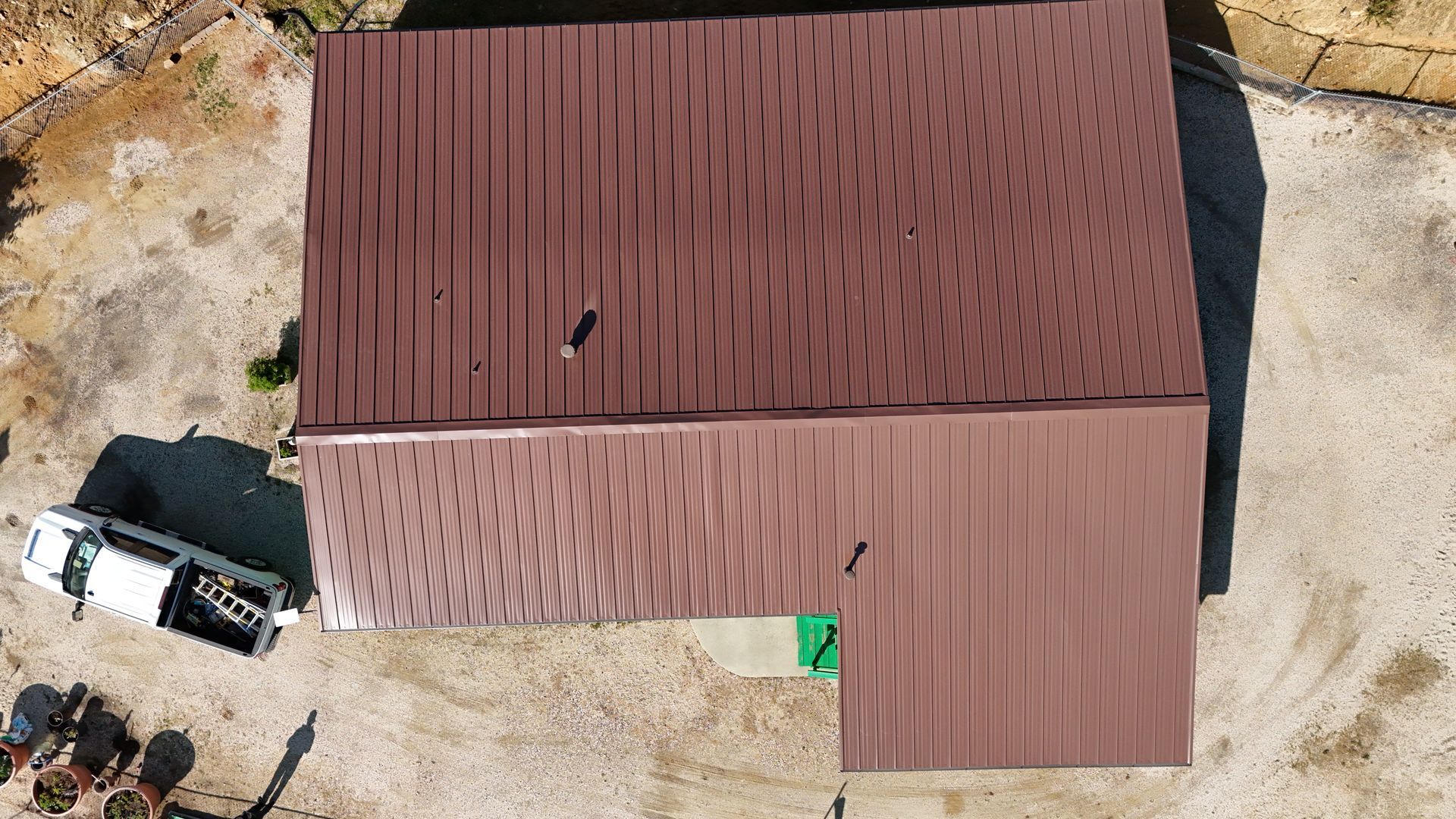 Aerial view of a brown-roofed building beside a white van on a dirt lot.