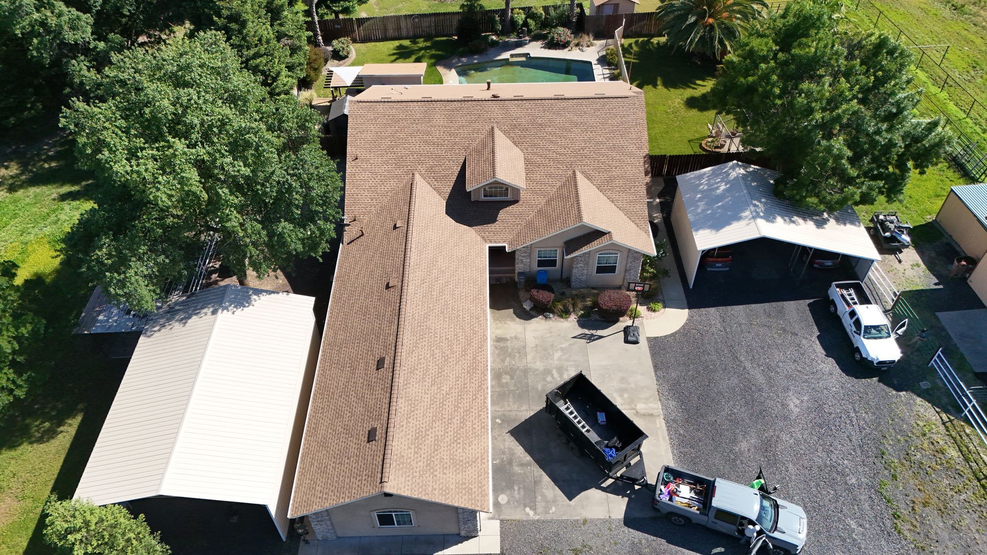 Aerial view of a brown-roofed house with a gravel driveway, carport, and parked vehicles surrounded by trees.