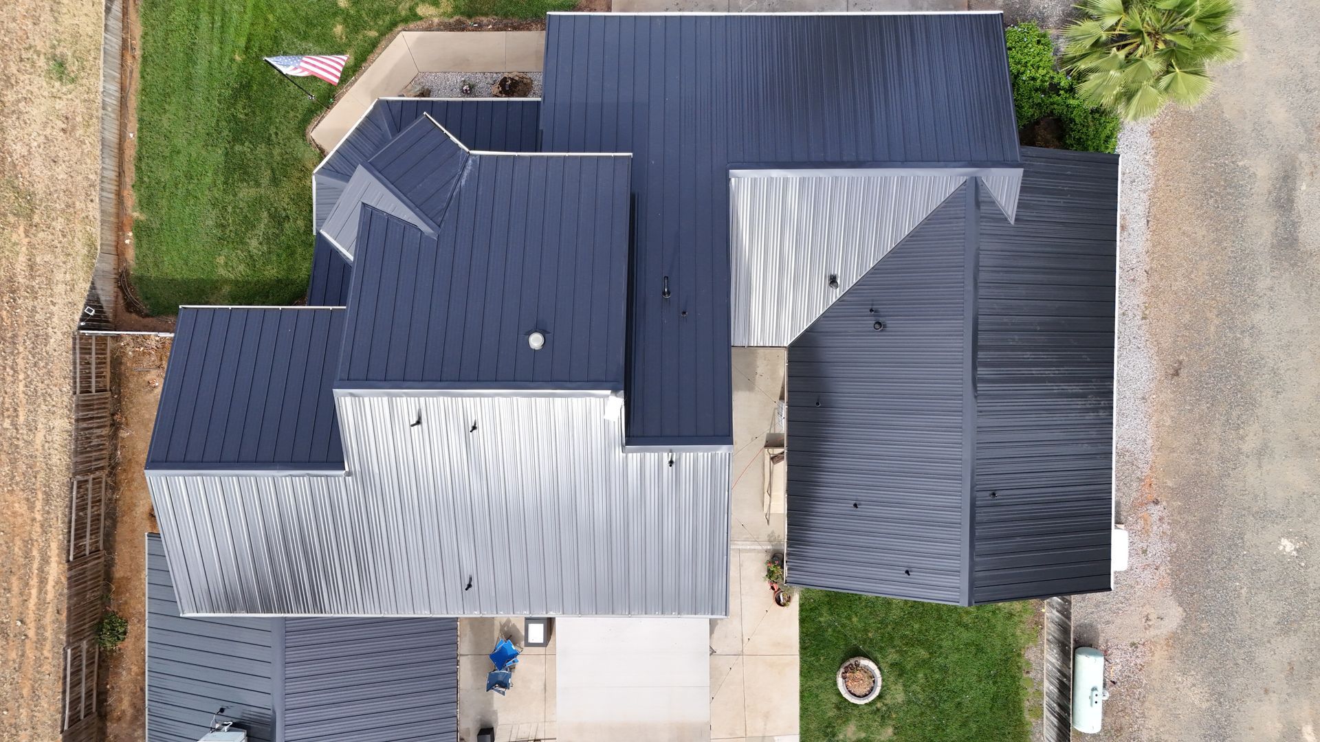 Aerial view of a house with a gray roof and driveway, surrounded by grass and neighboring yards.