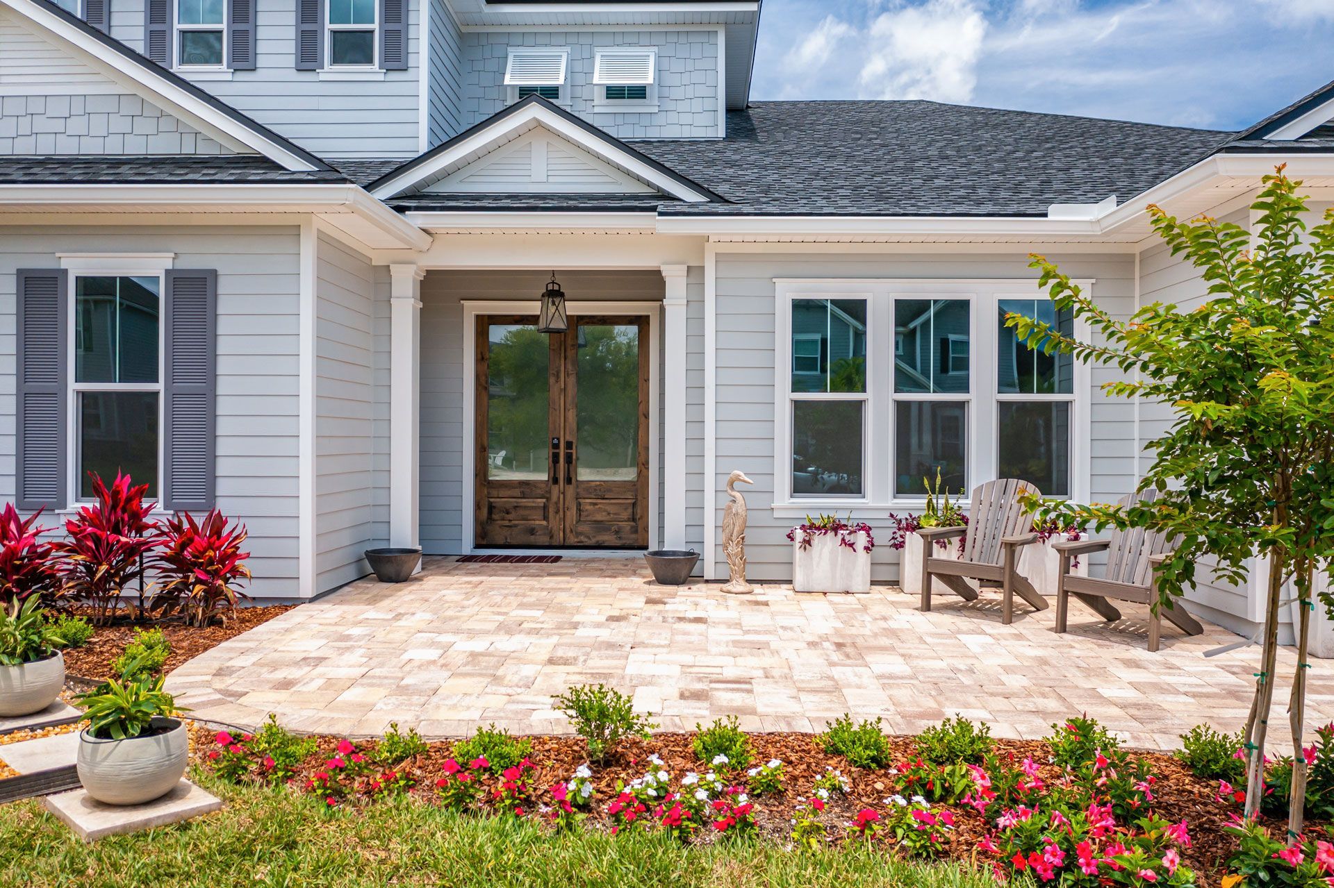 Front porch of a gray house with double doors, white chairs, and flower beds under a bright sky