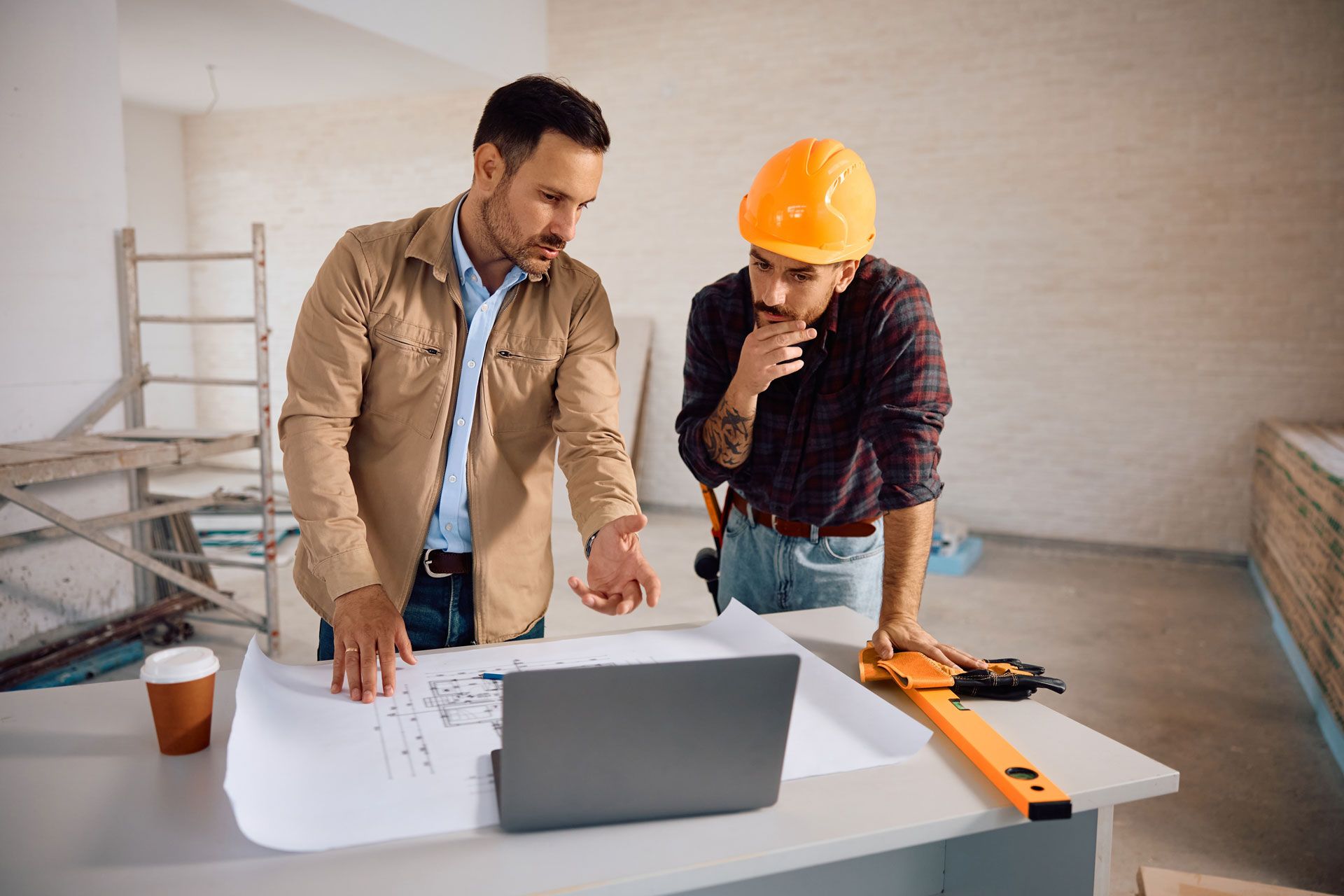 Two construction workers reviewing blueprints and plans at a table with a laptop in a renovation site