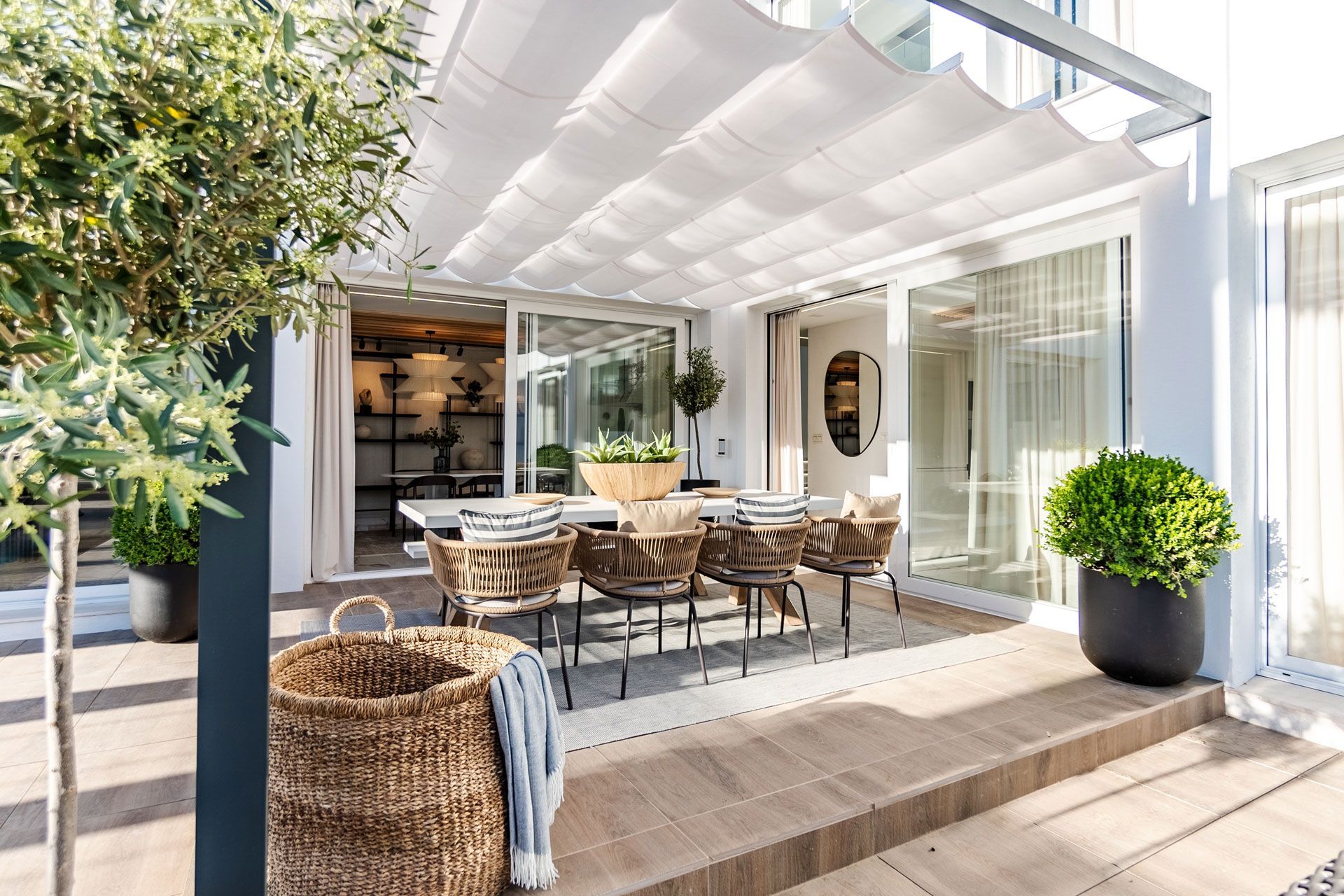 Modern patio dining area with wicker table and chairs under a white pergola, beside a bright house interior