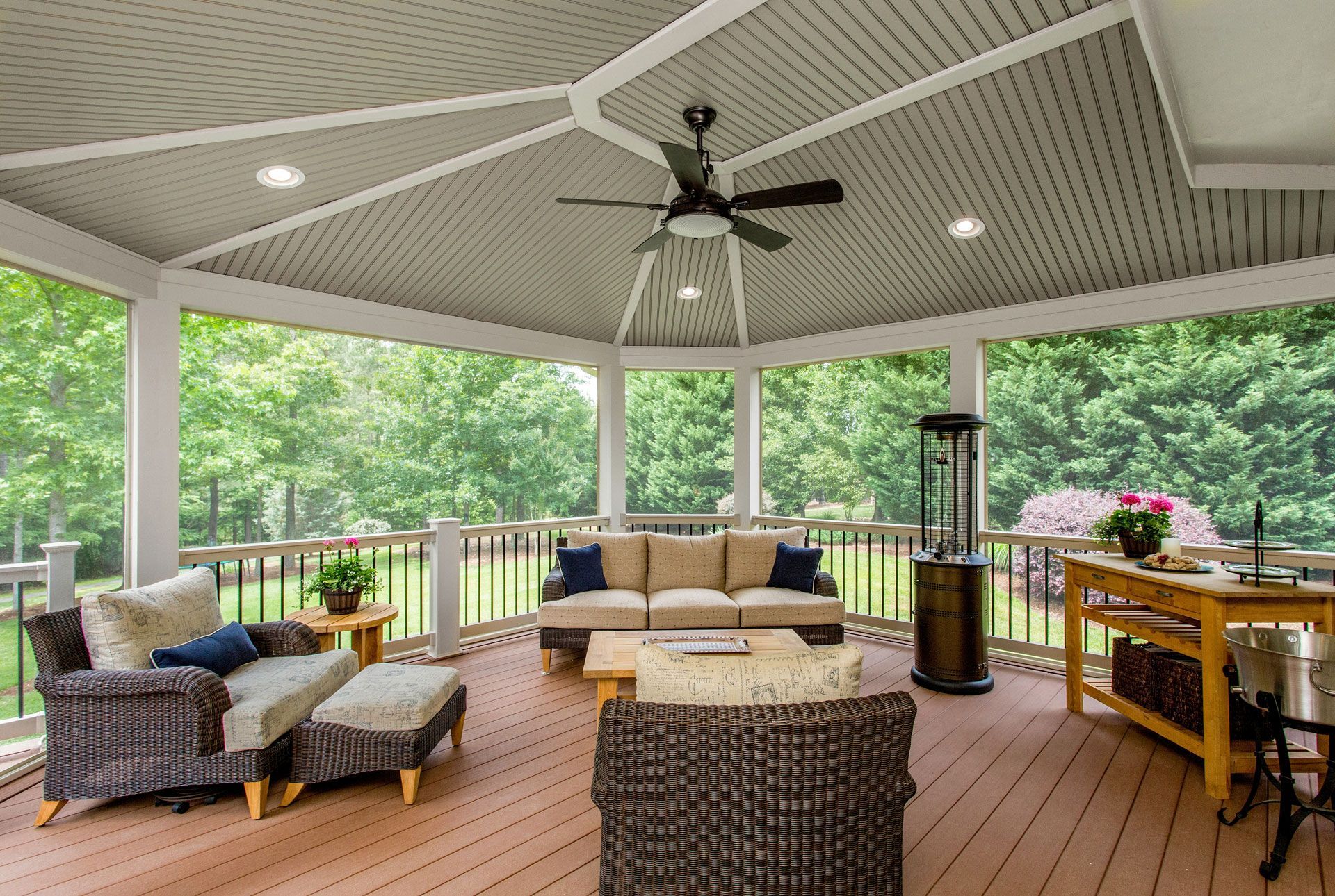 Covered outdoor deck with wicker seating, ceiling fan, grill, and wooden table overlooking green trees