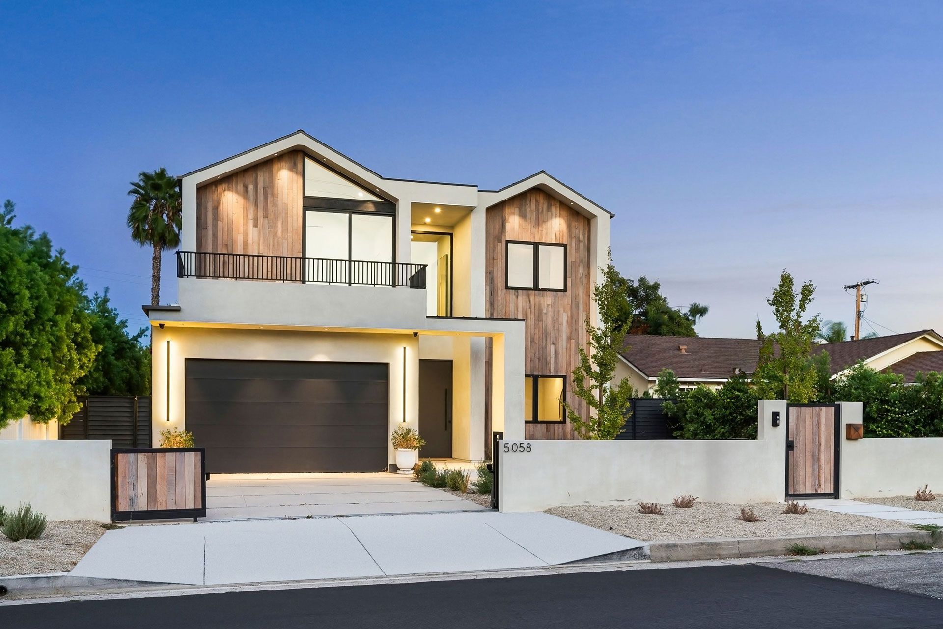 Modern two-story house with warm exterior lighting, garage, and white front wall at dusk